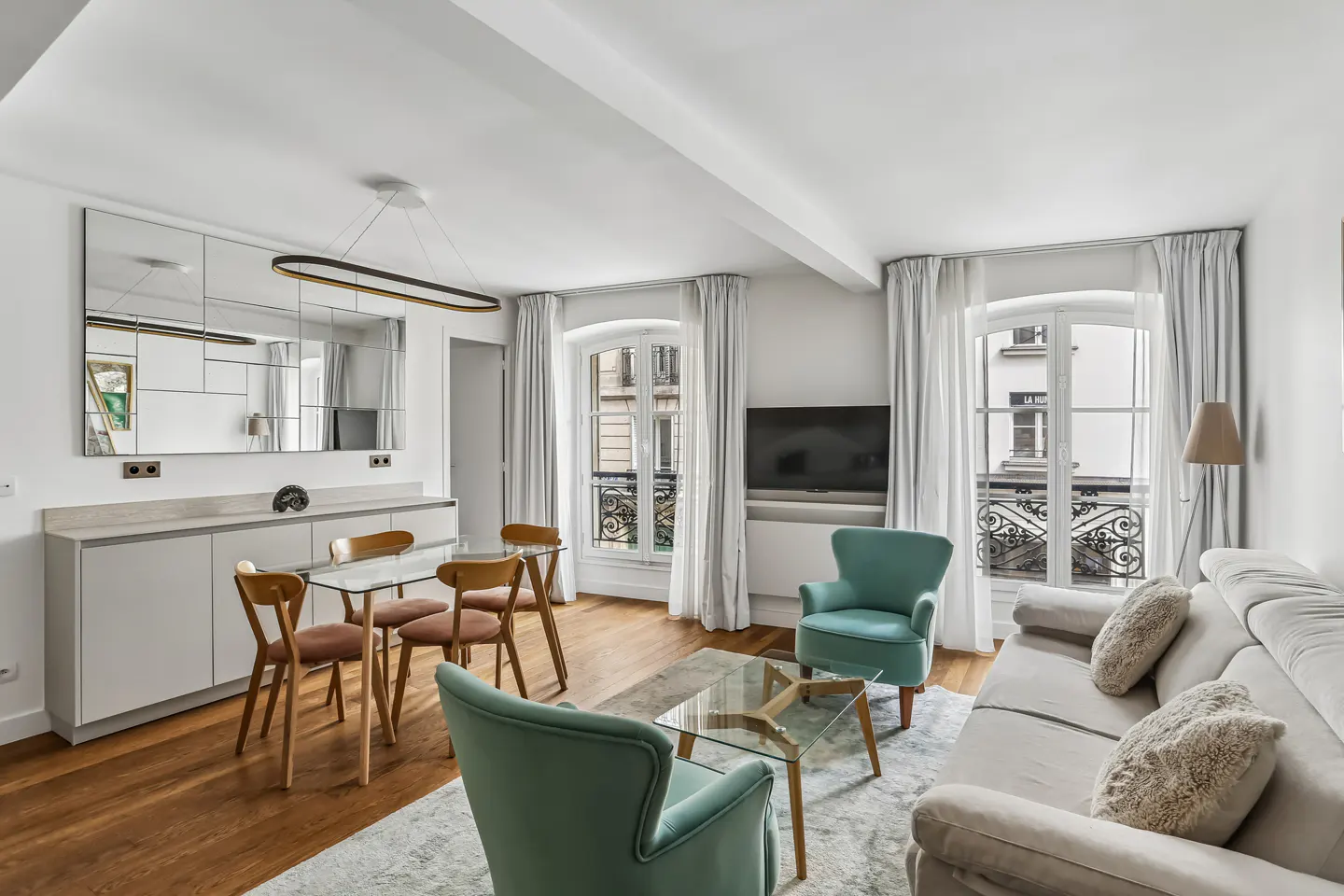 Bright living room with wood floors, white walls, and large windows. Features a dining table, sofa, armchairs, and a TV.
