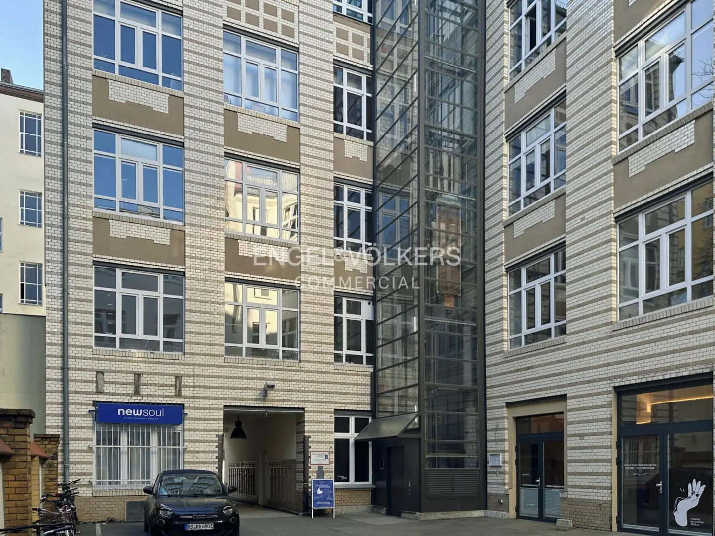 Exterior view of a multi-story brick building with large windows and a glass elevator shaft. A black car is parked in front.