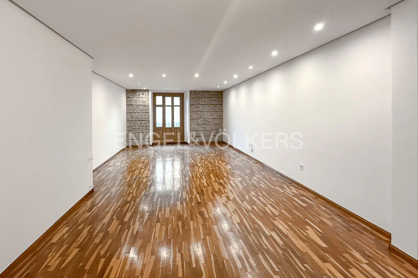 Empty room with hardwood floors, white walls, and a brick wall with a wooden door at the end.