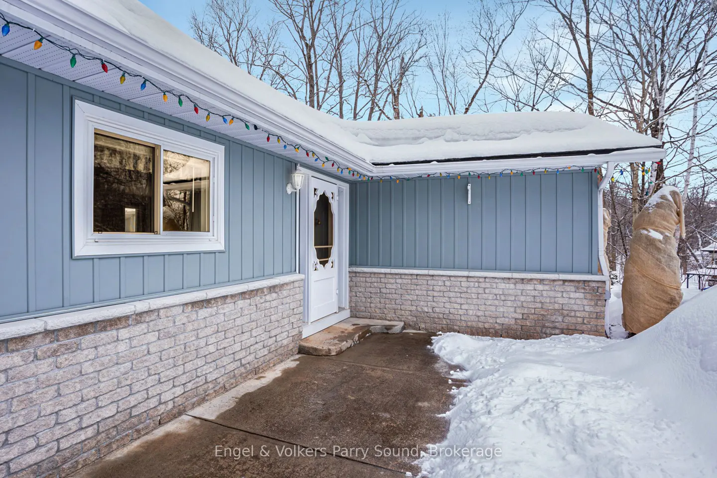 Exterior of a light blue house with white trim, brick foundation, and Christmas lights. Snow on the roof and ground.