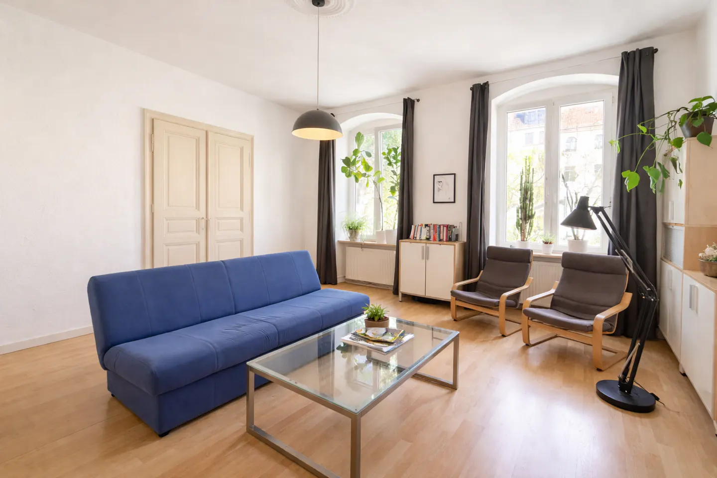 Bright living room with a blue sofa, glass table, two chairs, and large windows with gray curtains.