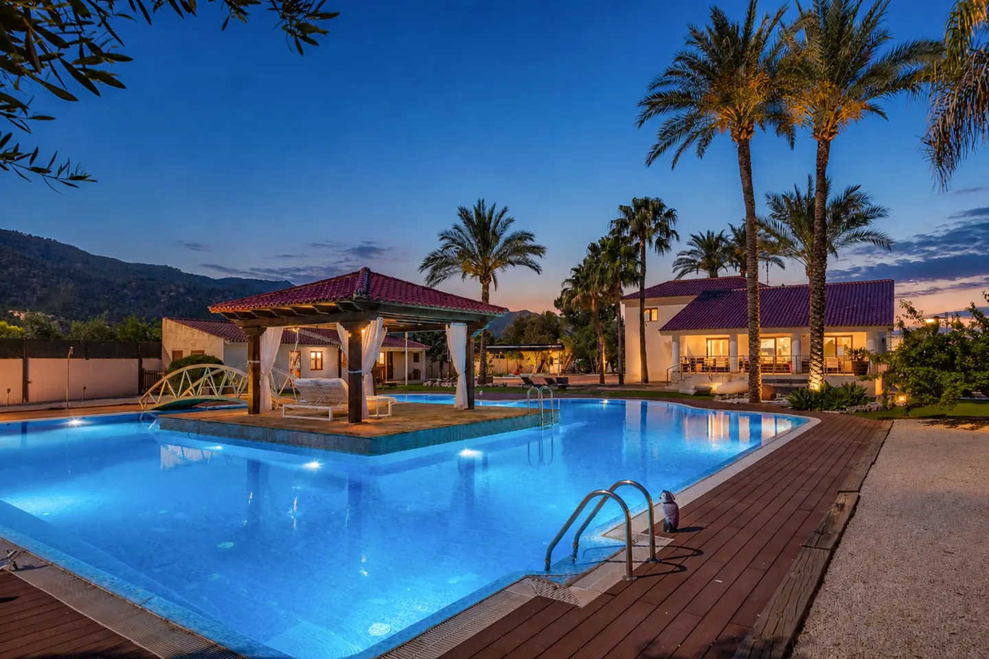 Luxury villa with a blue-lit pool, gazebo with white curtains, and palm trees against a twilight sky.