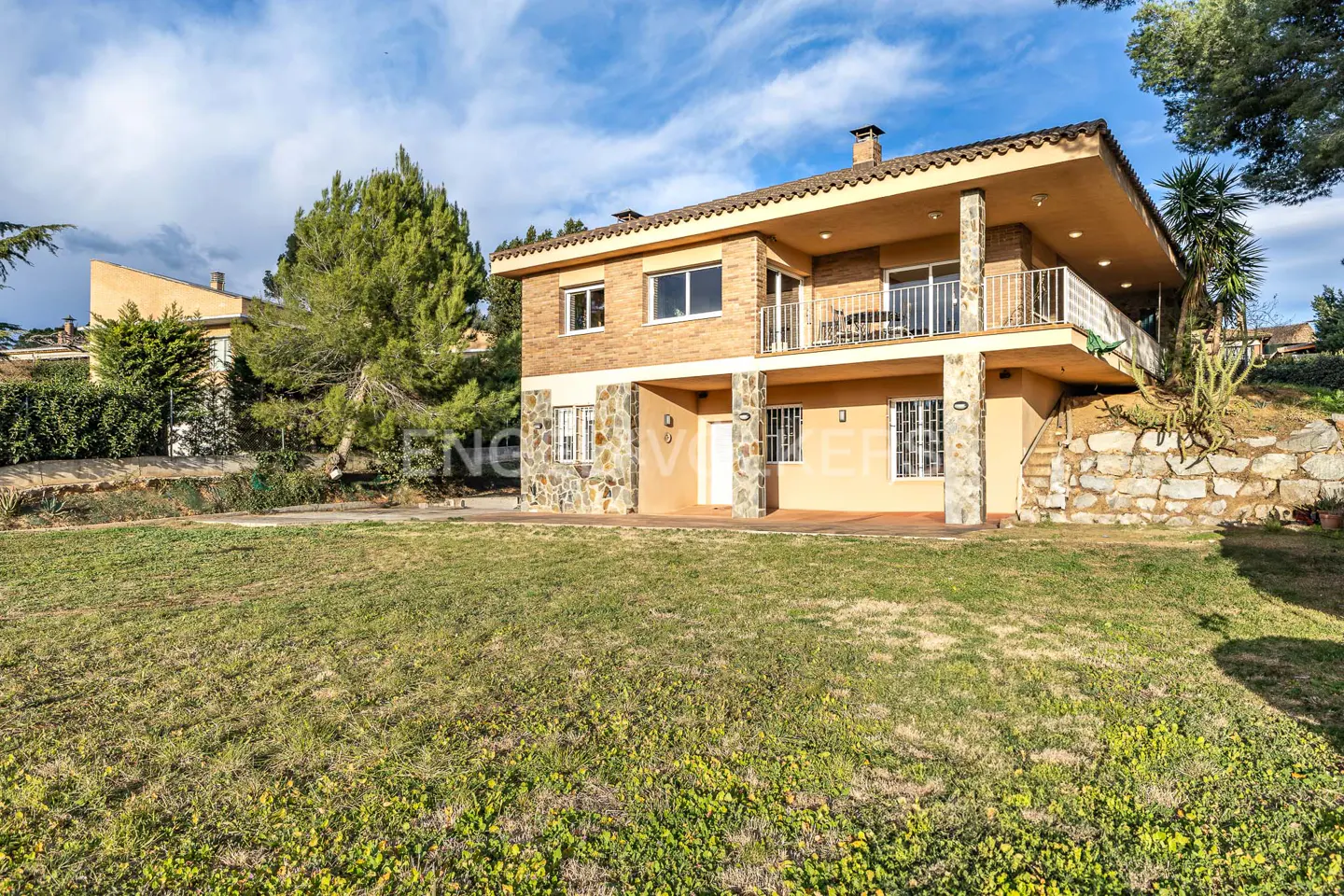 Two-story house with a balcony, stone accents, and a large lawn under a partly cloudy sky.