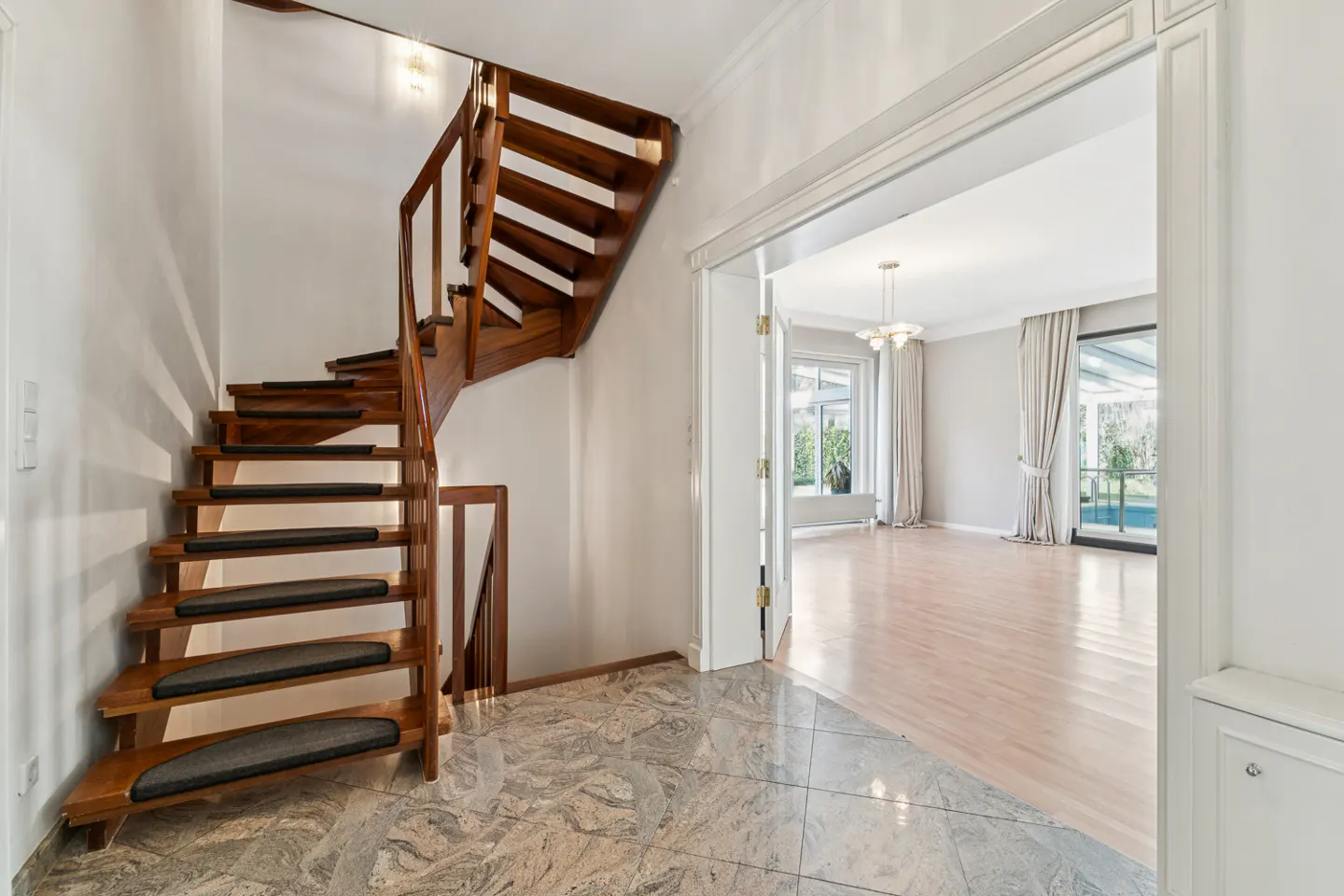 Entryway with a wooden staircase and marble floor. An open doorway leads to a bright living room with a chandelier and large windows.