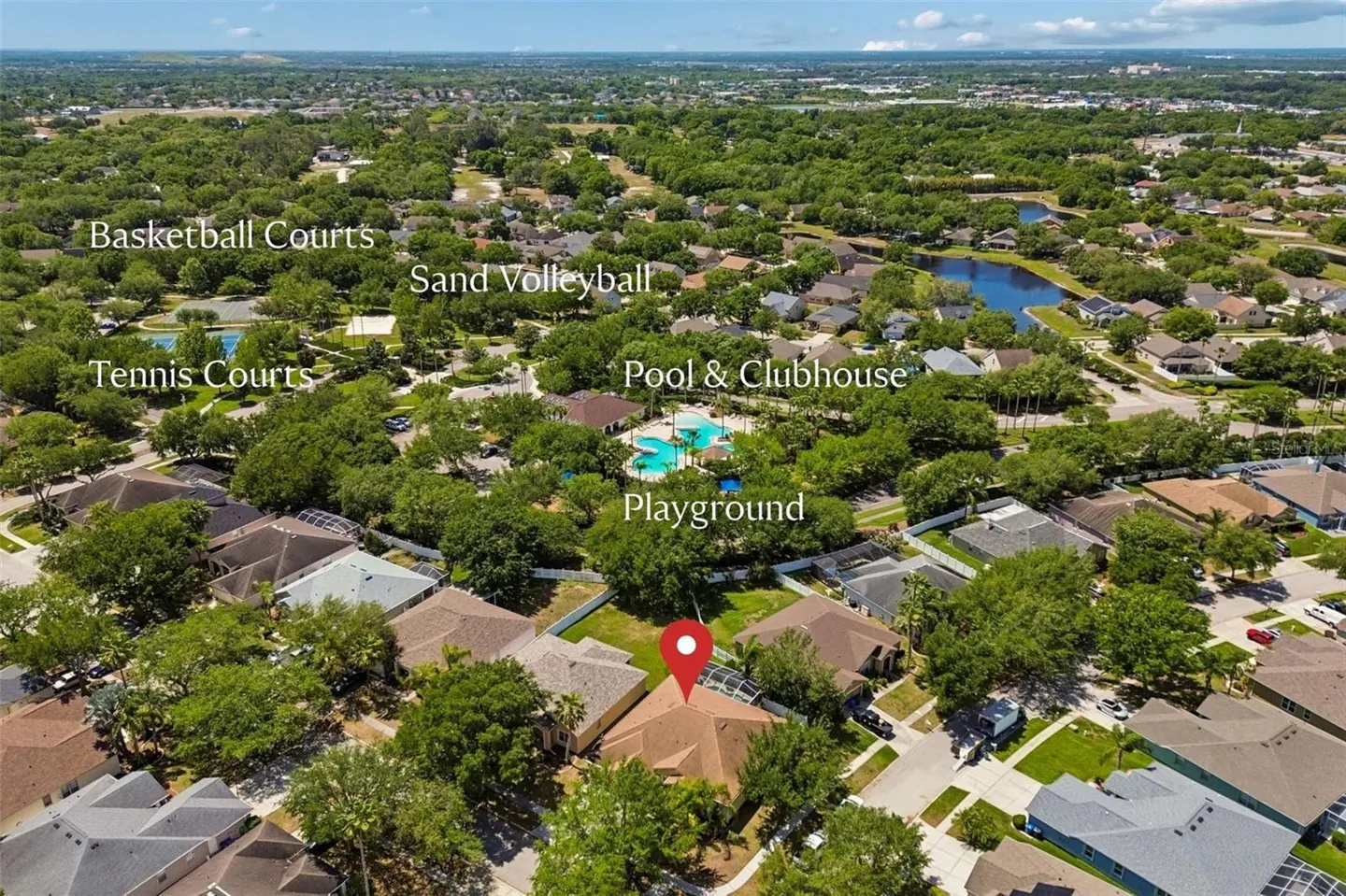 Aerial view of a residential neighborhood with amenities like a pool, playground, and sports courts. A red pin marks a house with a brown roof.