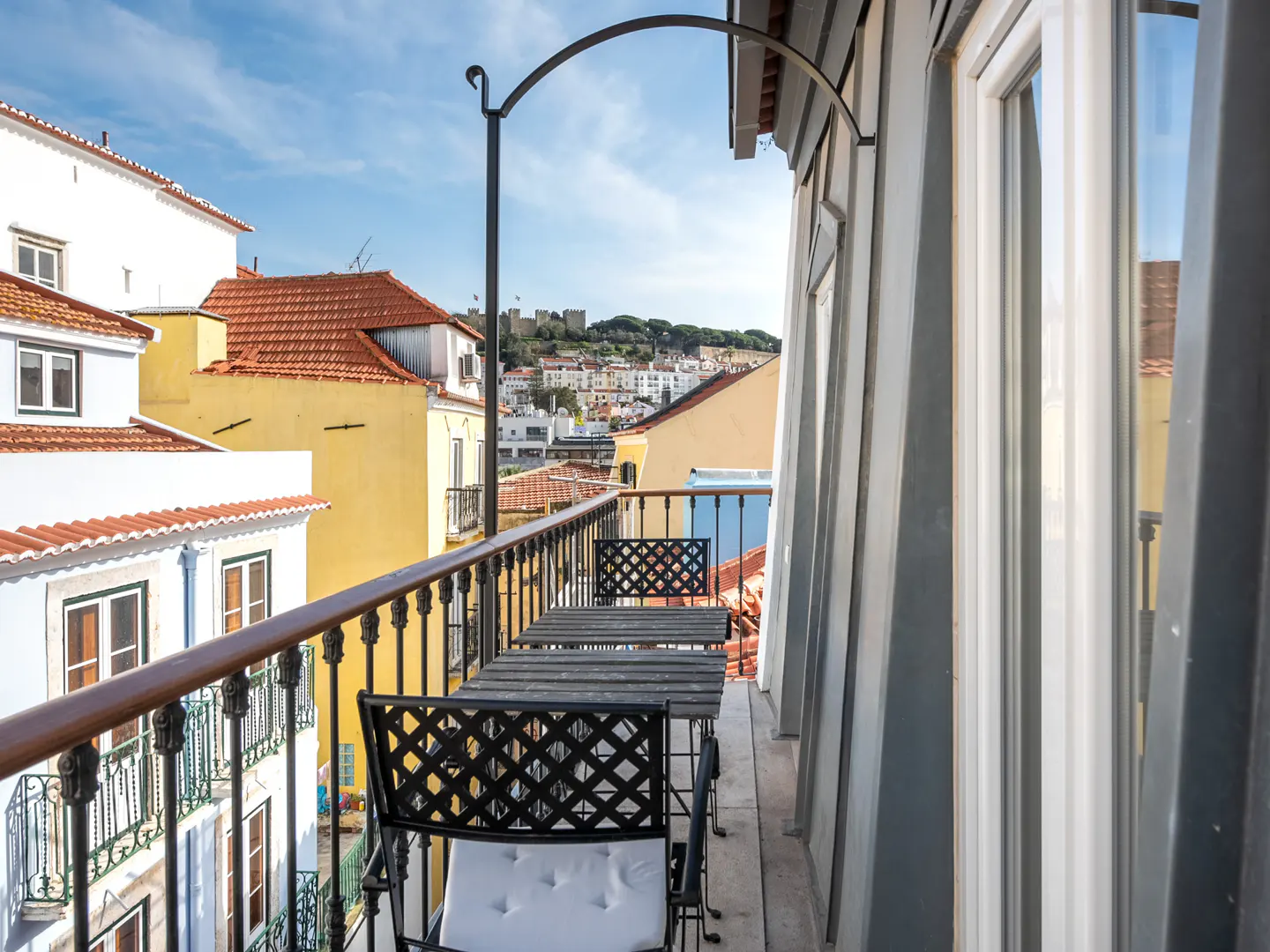 Balcony view with black metal table and chairs overlooking colorful buildings and a distant castle under a blue sky.