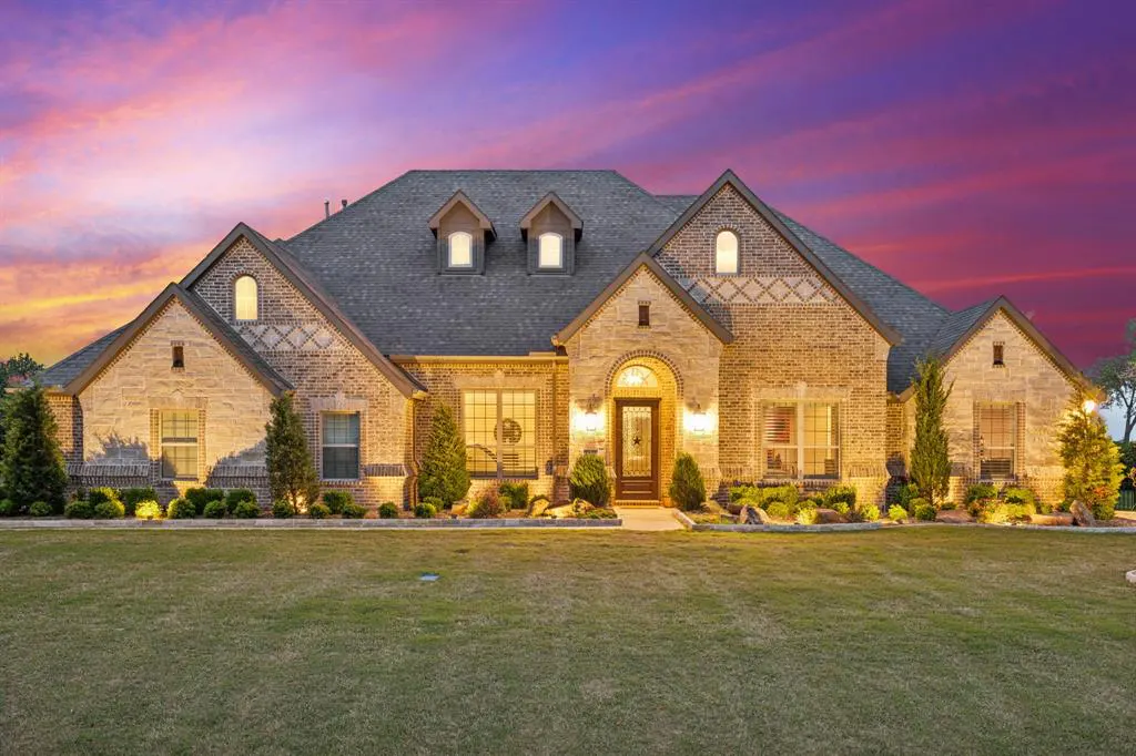 A tan brick house with a gray roof sits on a green lawn under a colorful sunset sky.