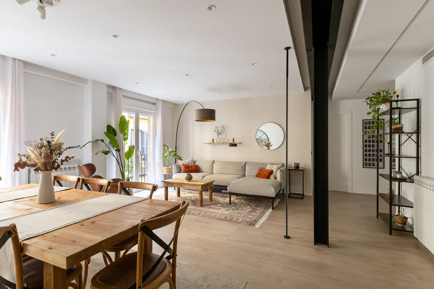 Open-concept living space with a wooden dining table, beige sofa, and black shelving unit. Natural light fills the room.