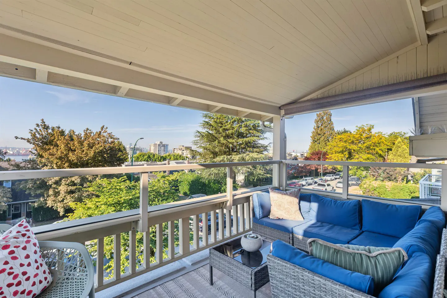 Covered balcony with blue cushioned wicker furniture, a glass railing, and a city view with trees.