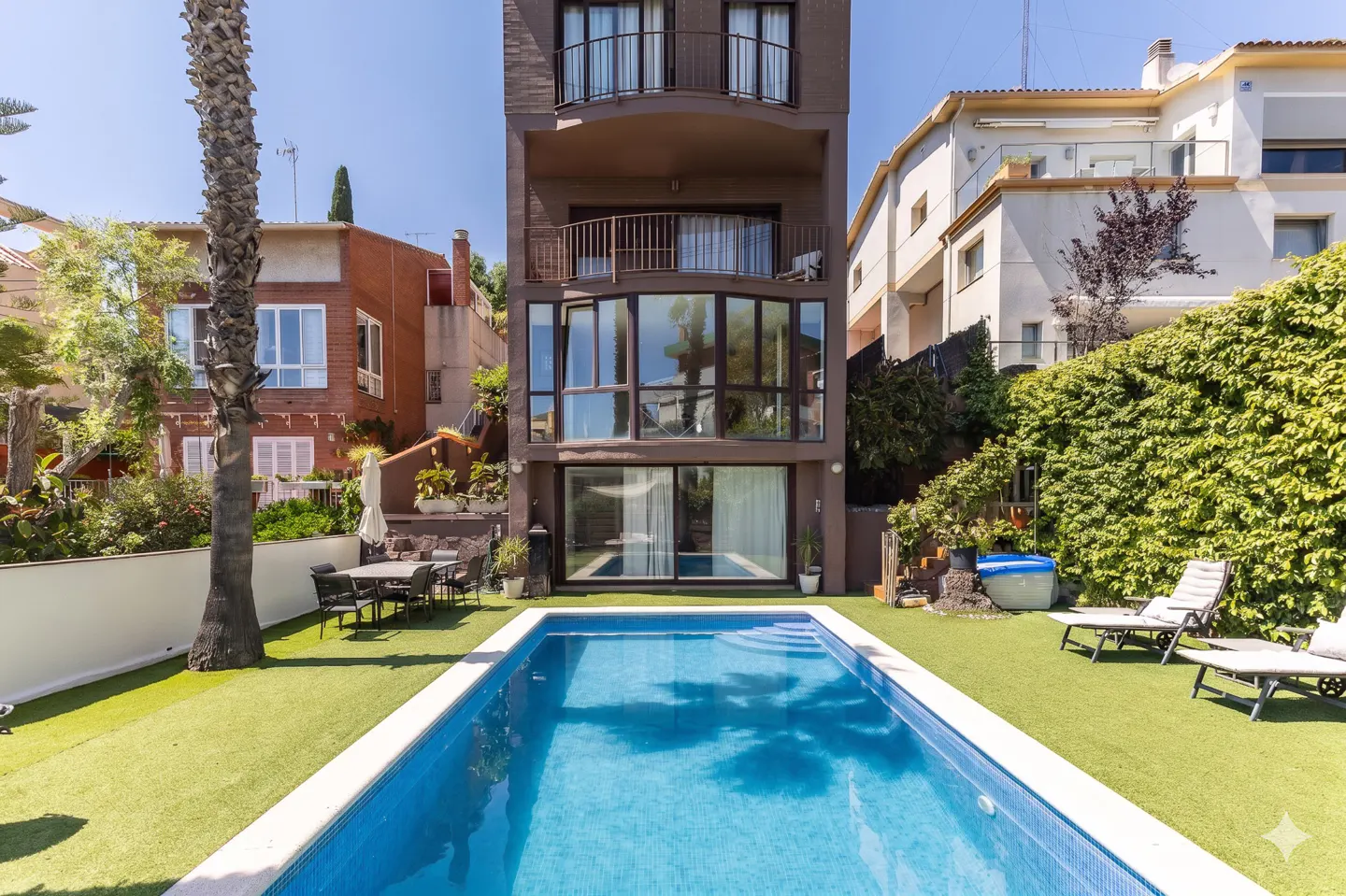 A modern brown three-story house with a blue tiled pool and green lawn. Outdoor furniture and lush greenery surround the property.
