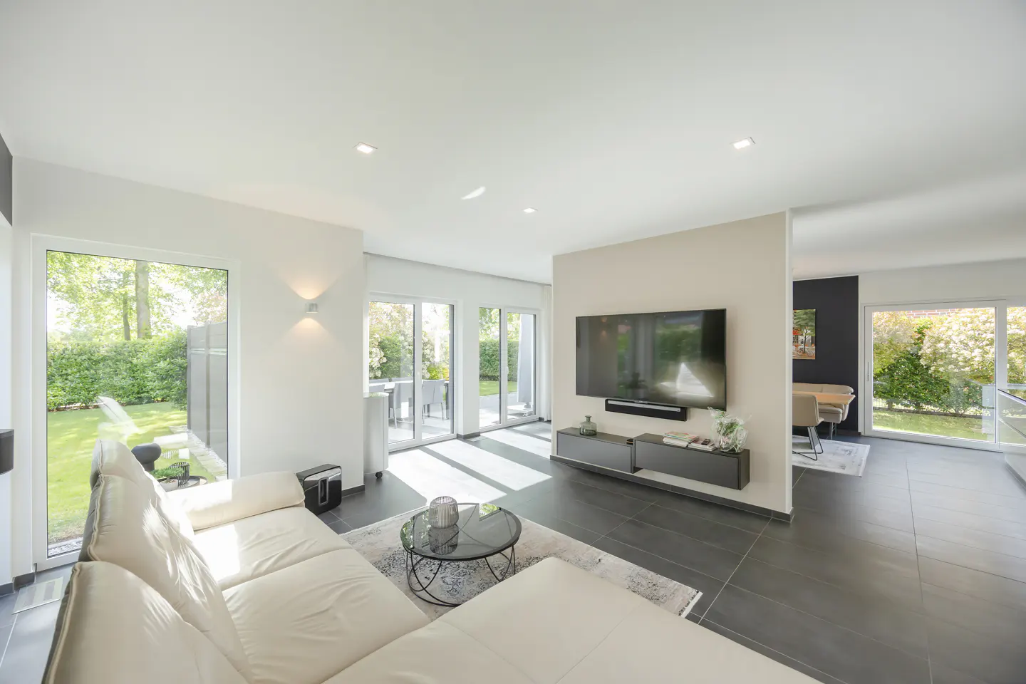 Bright, modern living room with white sofa, glass table, and large TV. Sliding glass doors lead to a green garden.