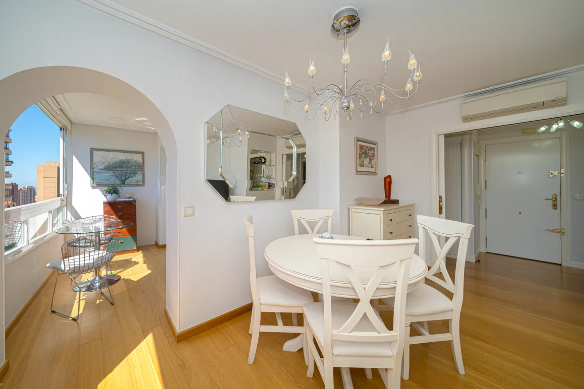 Bright dining area with white round table, chairs, and chandelier. Balcony with city view through arched doorway. Hardwood floors.