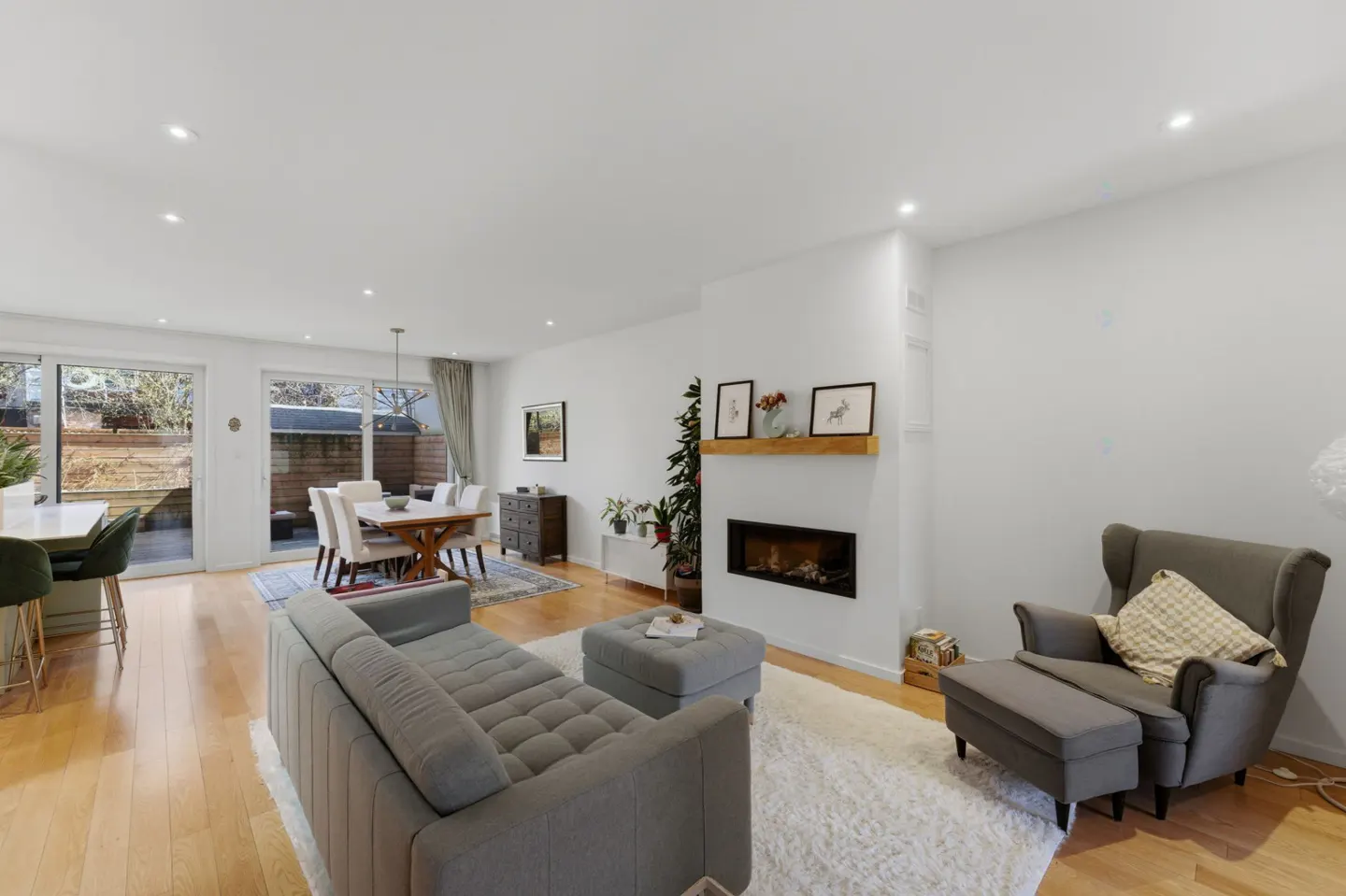 Bright, open-concept living room with hardwood floors, gray sofa, fireplace, dining table, and large windows.