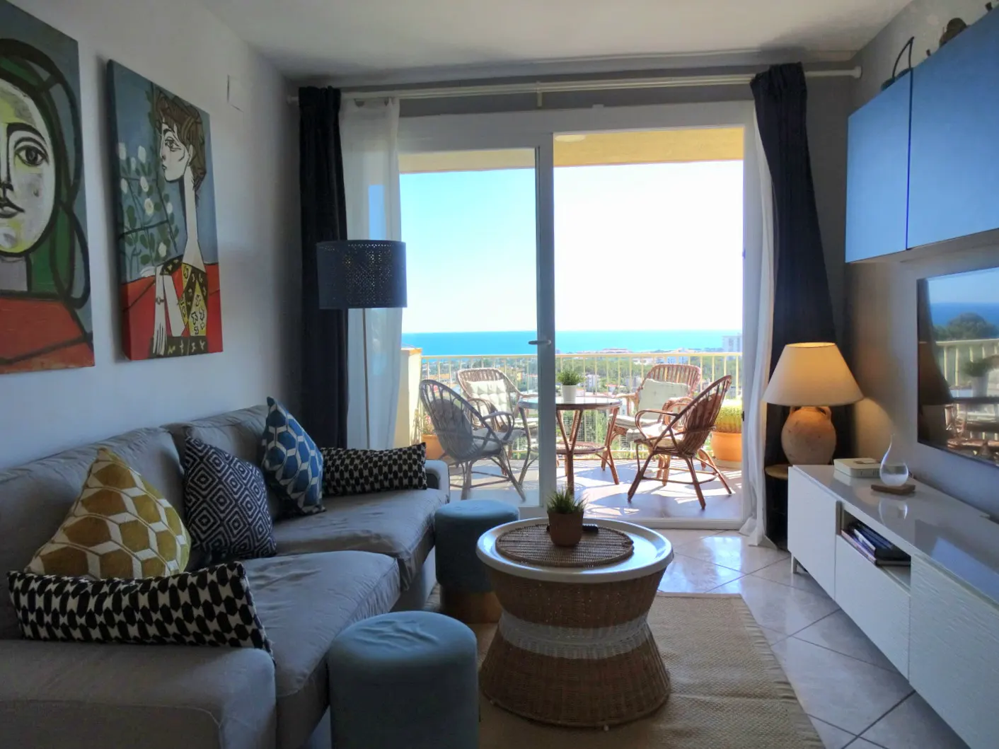 Living room with gray sofa, blue accents, and balcony overlooking the ocean. Picasso-style art on the wall.