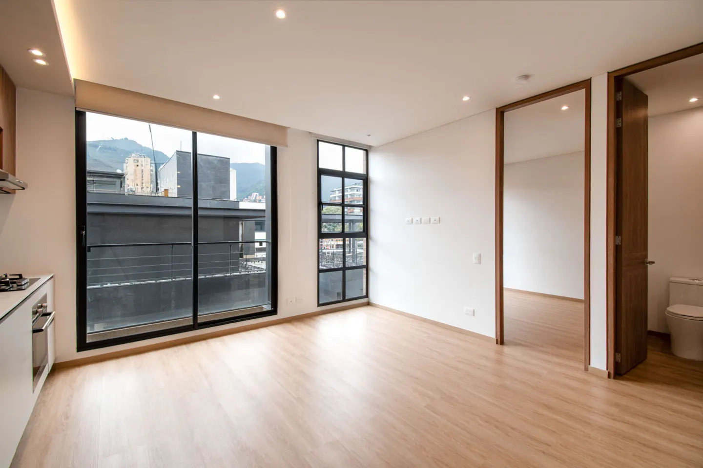Bright, empty apartment with wood floors, white walls, and large windows with city views. A bathroom is visible through an open door.
