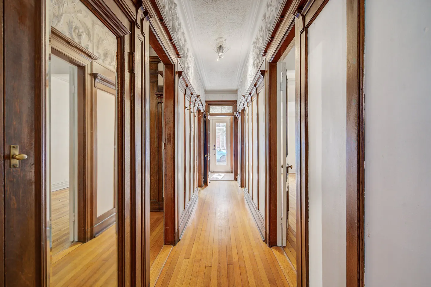 Long hallway with wood floors, white walls, and dark wood trim. Doors line the hallway, leading to other rooms. A glass door is at the end.