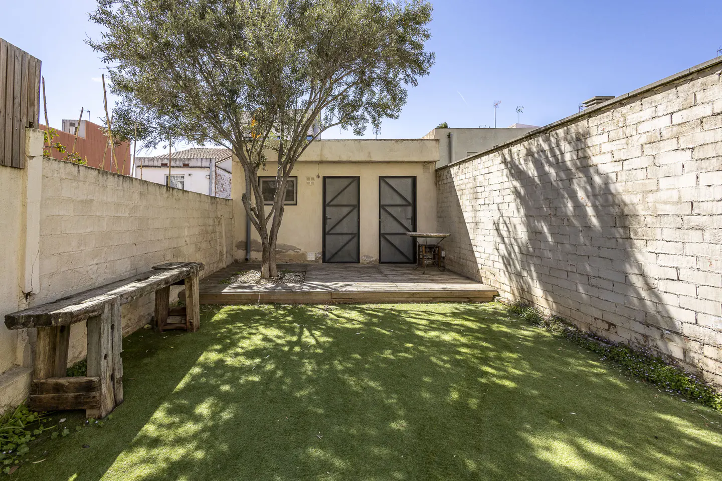A backyard with green grass, a tree, a wooden bench, and a small building with two doors. The yard is surrounded by white brick walls.