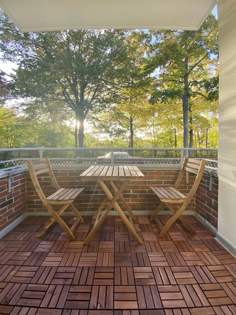 Balcony with wood tile flooring, brick wall, and a folding wood table and chairs overlooking trees.