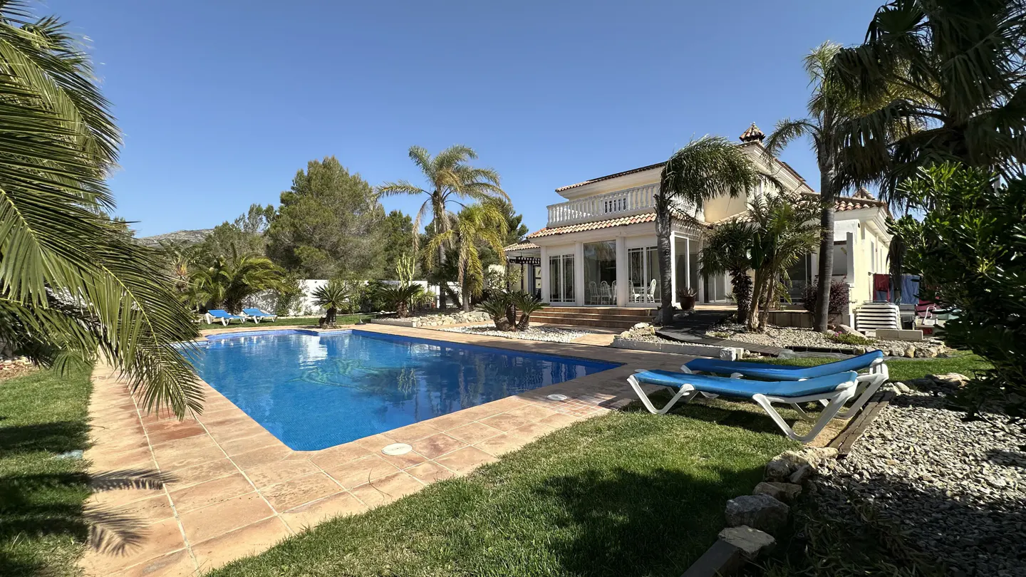 A sunny backyard with a blue pool, lounge chairs, palm trees, and a beige house with a balcony.