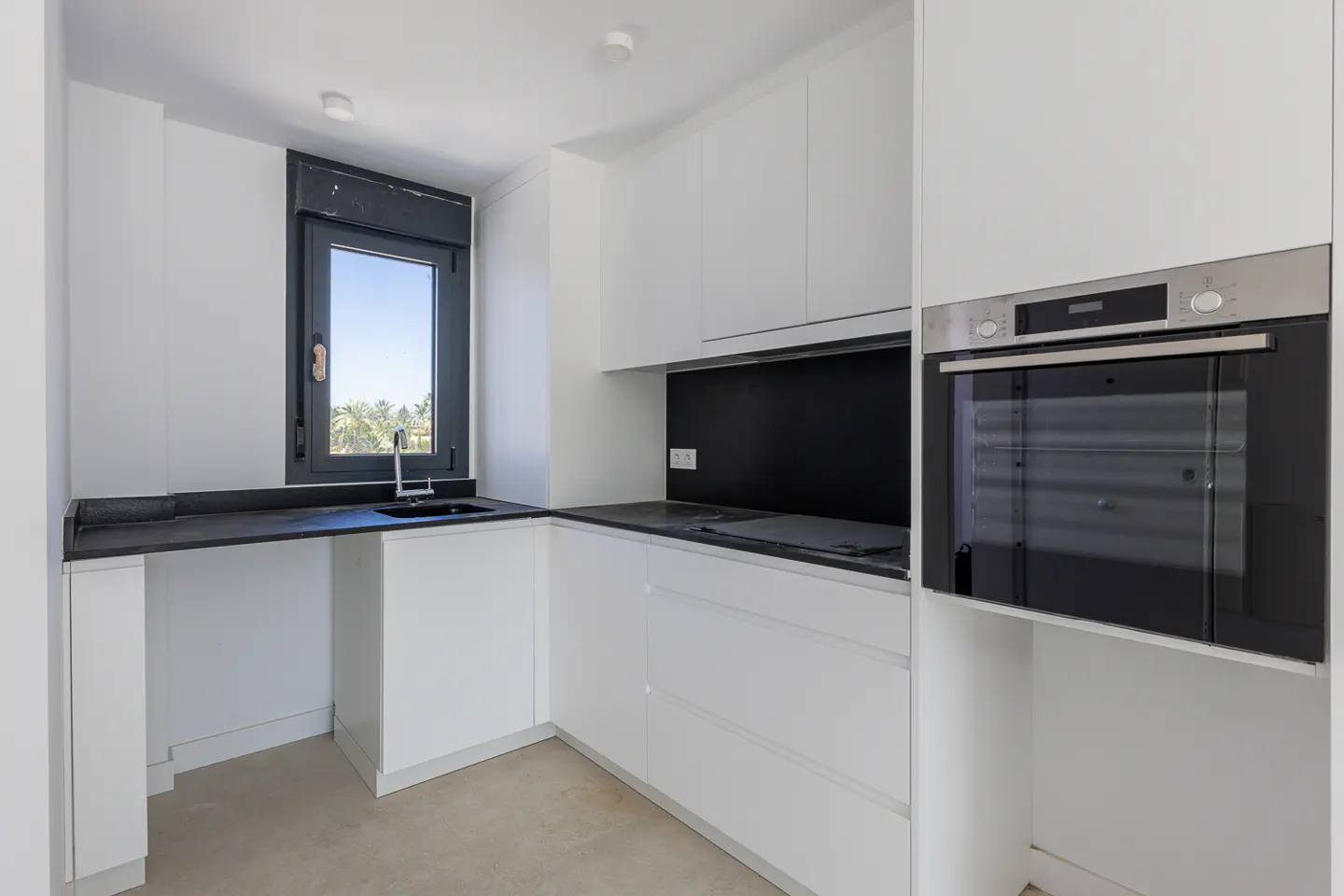 Modern kitchen with white cabinets, black countertops, a built-in oven, and a window overlooking a sunny outdoor area.