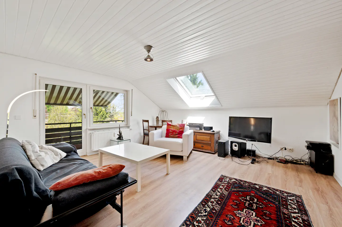 Attic living room with a black sofa, white chair, TV, and a red patterned rug on a light wood floor.