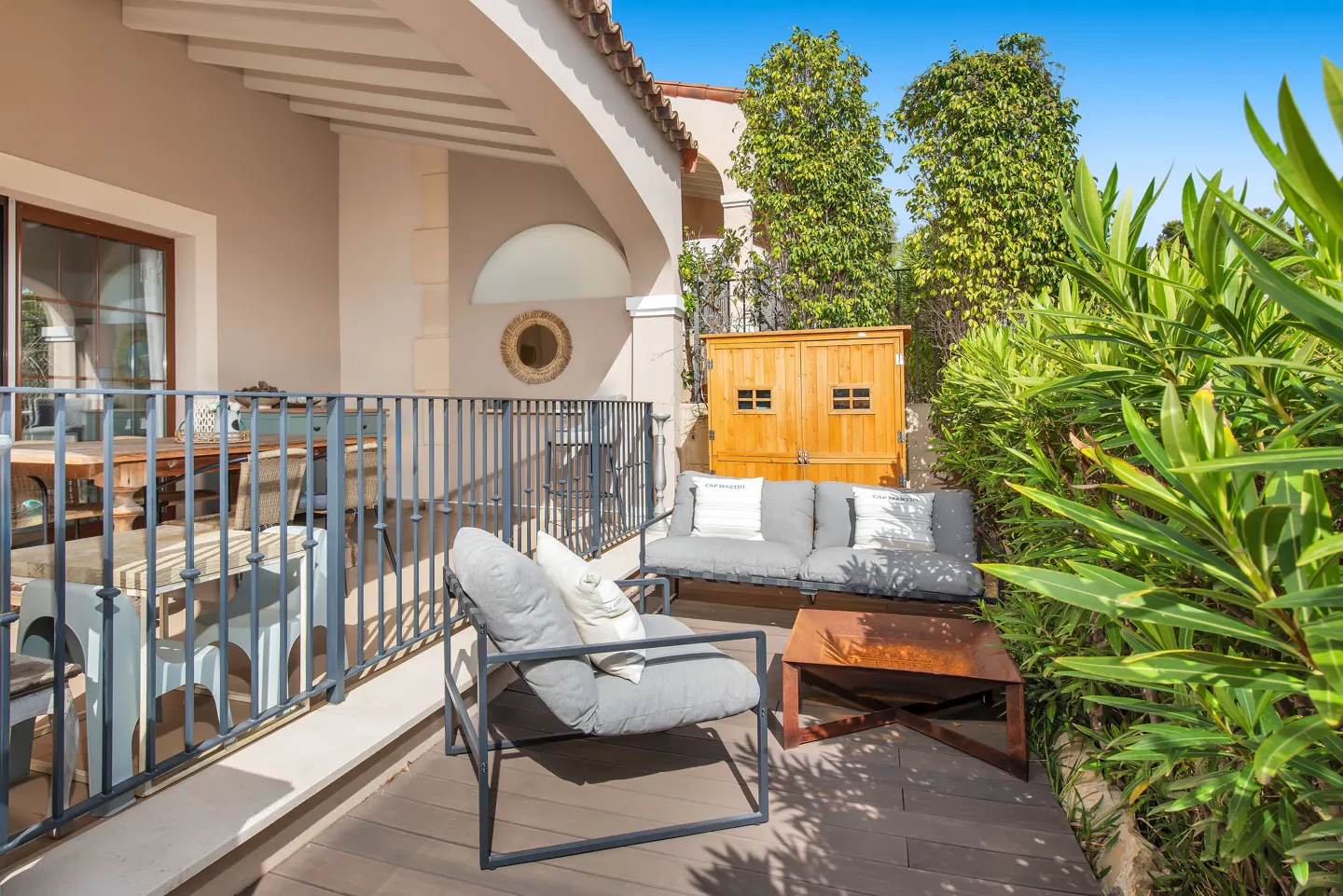A patio with gray furniture, a wooden cabinet, and lush greenery under a blue sky.