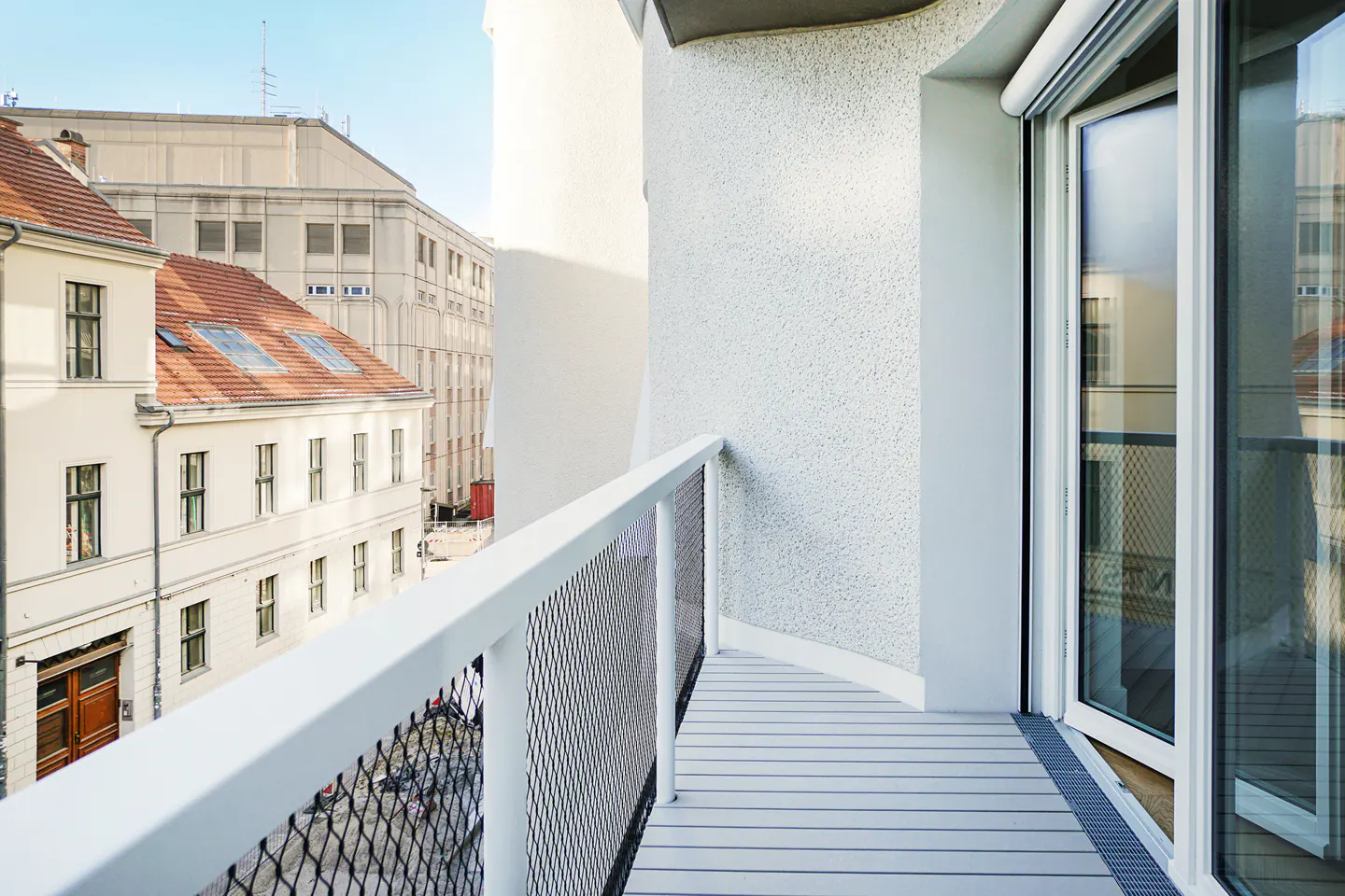 View from a white balcony with a metal railing, overlooking buildings with red roofs on a sunny day.