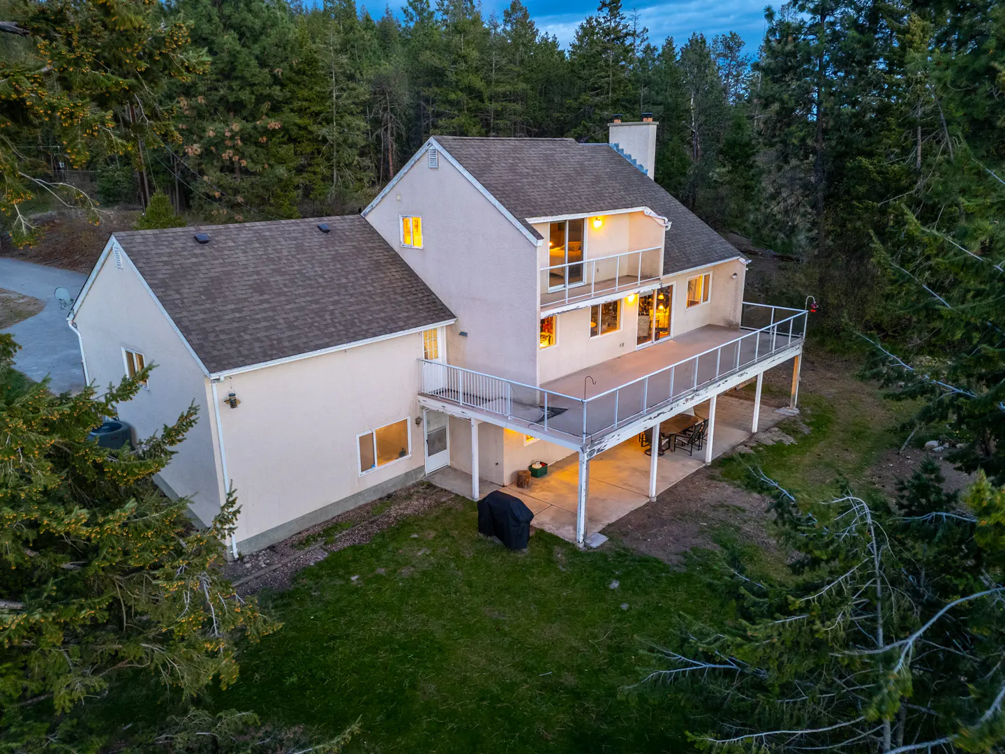 Two-story beige house with a large deck and a brown roof, surrounded by green trees and grass.