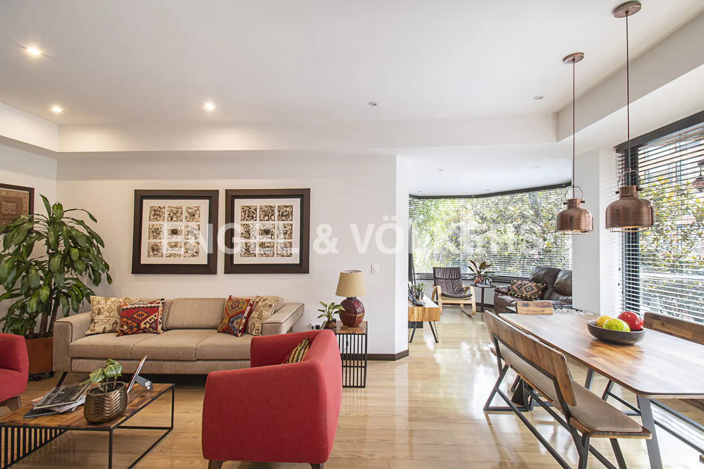 Bright living room with a beige sofa, red armchairs, wood floors, and a dining table with copper pendant lights.