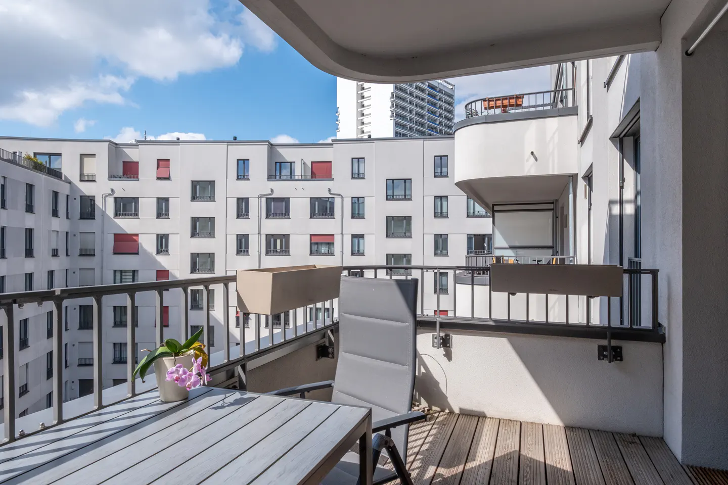 Balcony view with a table, chair, and potted orchid. White buildings and blue sky are in the background.