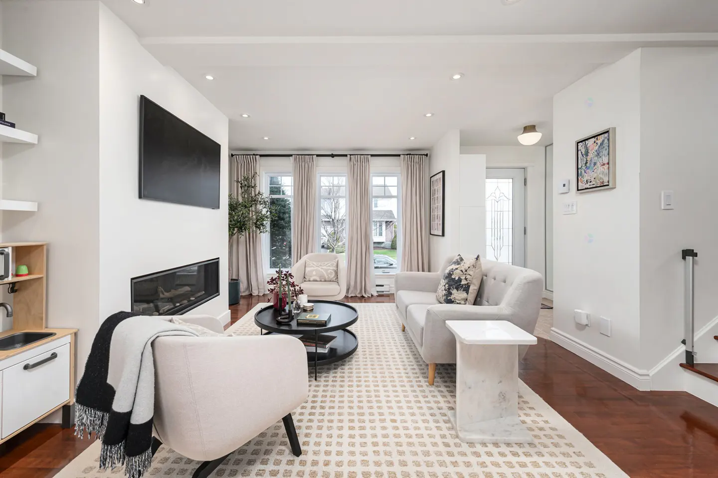 Bright living room with white walls, hardwood floors, and a patterned rug. A fireplace sits below a large TV, with sofas and chairs arranged around a black coffee table.