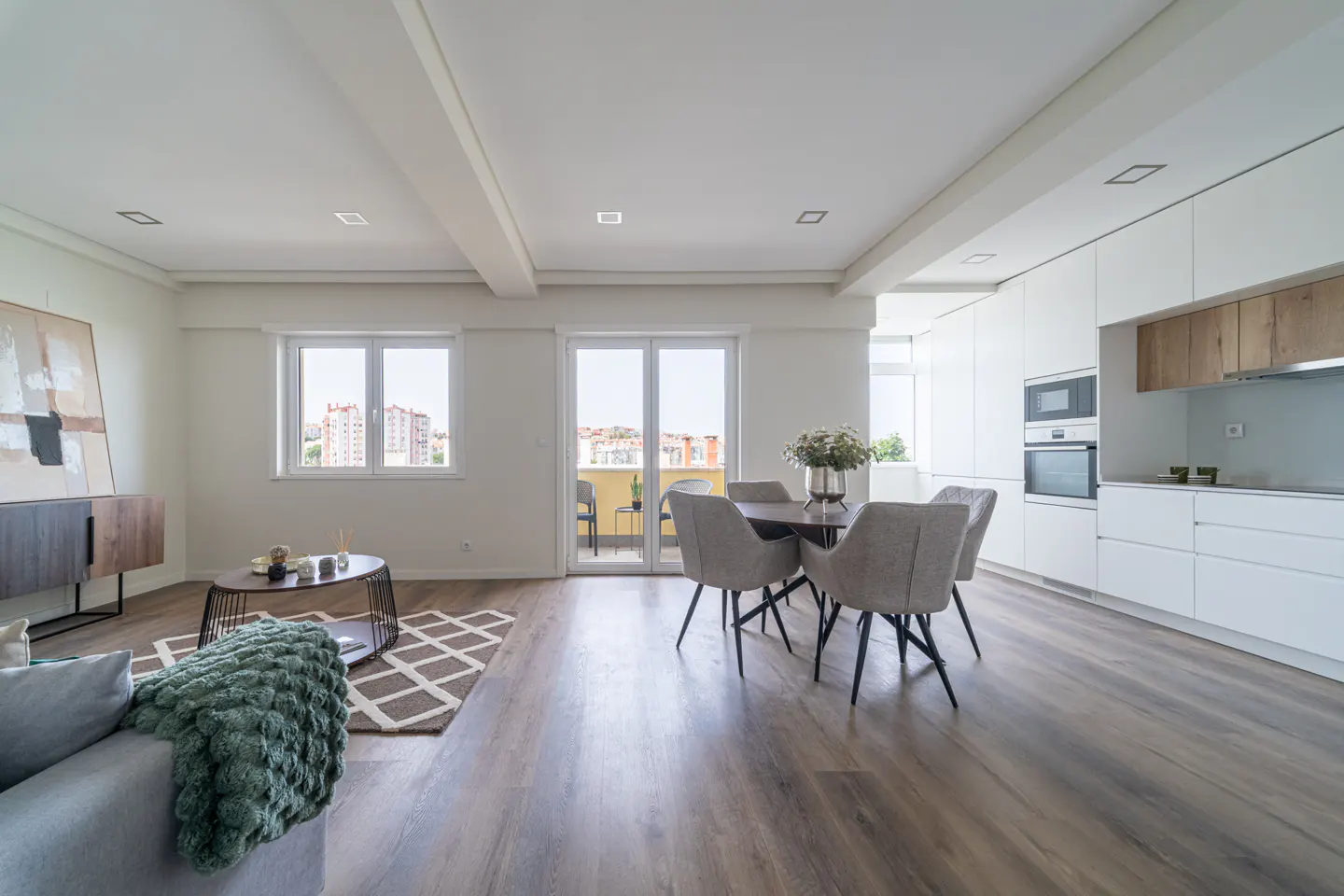 Bright, open-concept apartment with wood floors. Living room with gray sofa and green blanket. Dining area with round table and gray chairs. White kitchen cabinets. Balcony view.