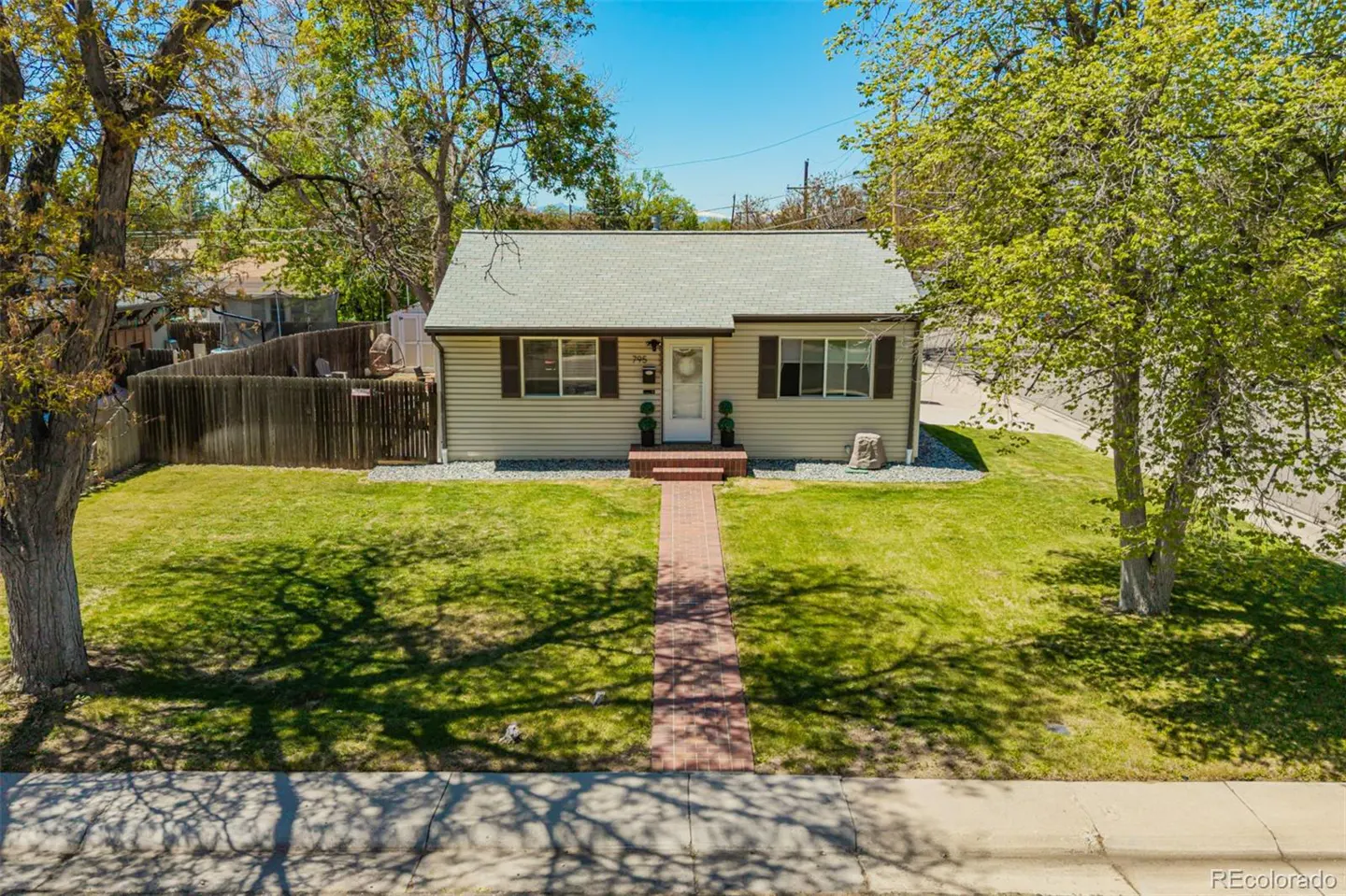 Tan single-story house with brown shutters and a brick walkway leading to a white front door, surrounded by green lawn and trees.