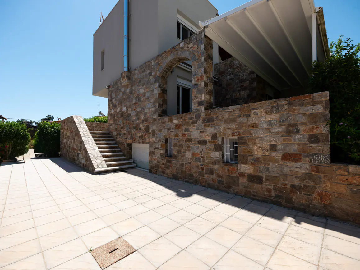 Modern house exterior with stone facade, tiled patio, and stone stairs leading to a covered terrace under a white awning.