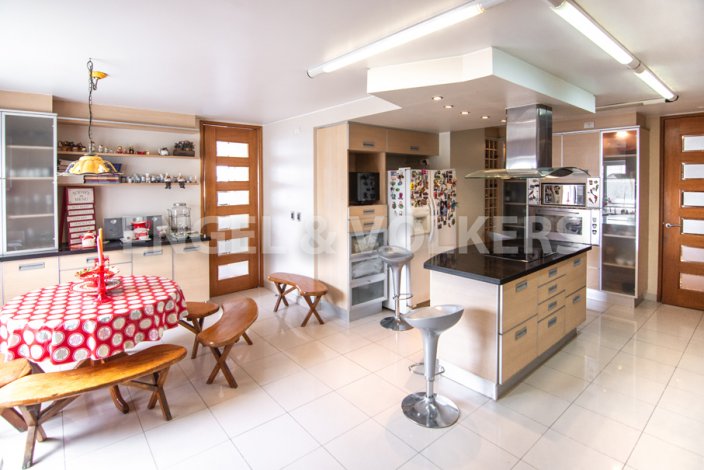 Open-concept kitchen with tile floors, light wood cabinets, and a black countertop island with silver stools. A round table with a red tablecloth and wooden benches are nearby. [AI-generated text]