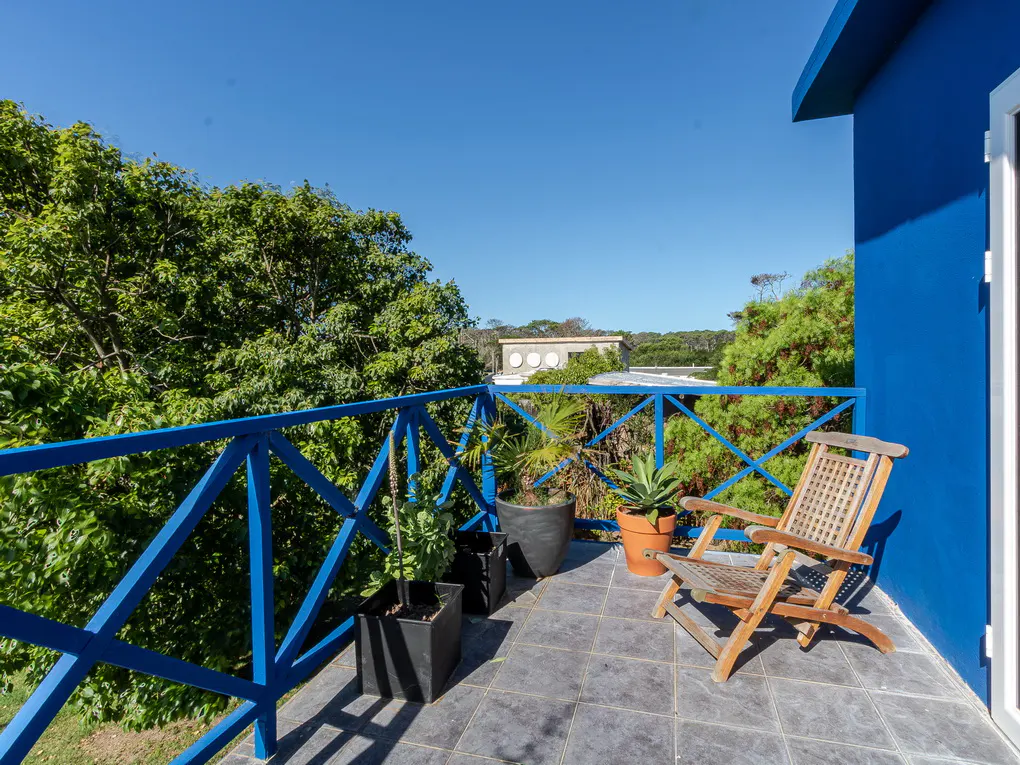 Balcony view with blue railings, potted plants, and a wooden chair. Lush green trees and a clear blue sky in the background.