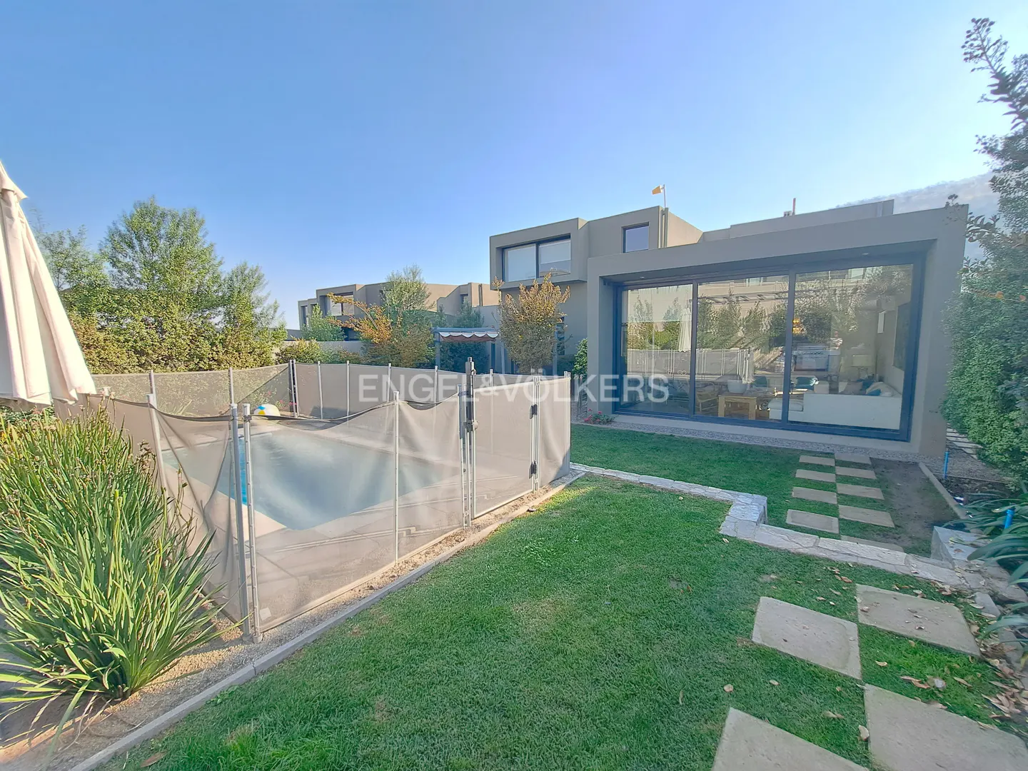 Exterior view of a modern, gray house with a pool and green lawn under a blue sky. A fence surrounds the pool area.