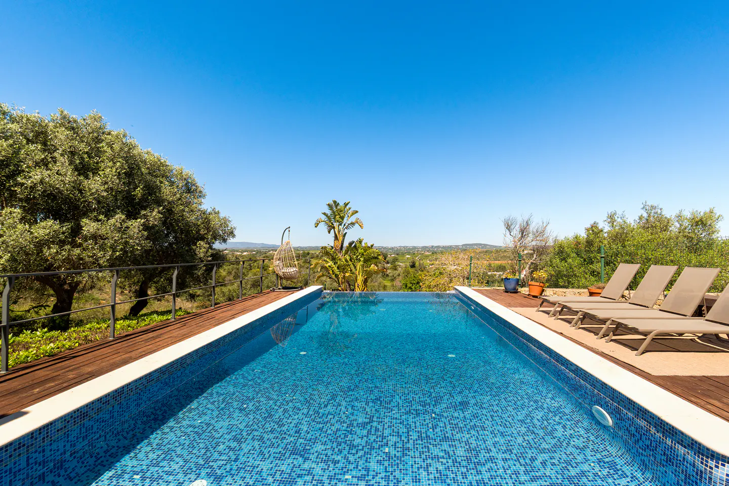 A blue tiled pool with lounge chairs and a hanging chair overlooks a green landscape under a clear blue sky.