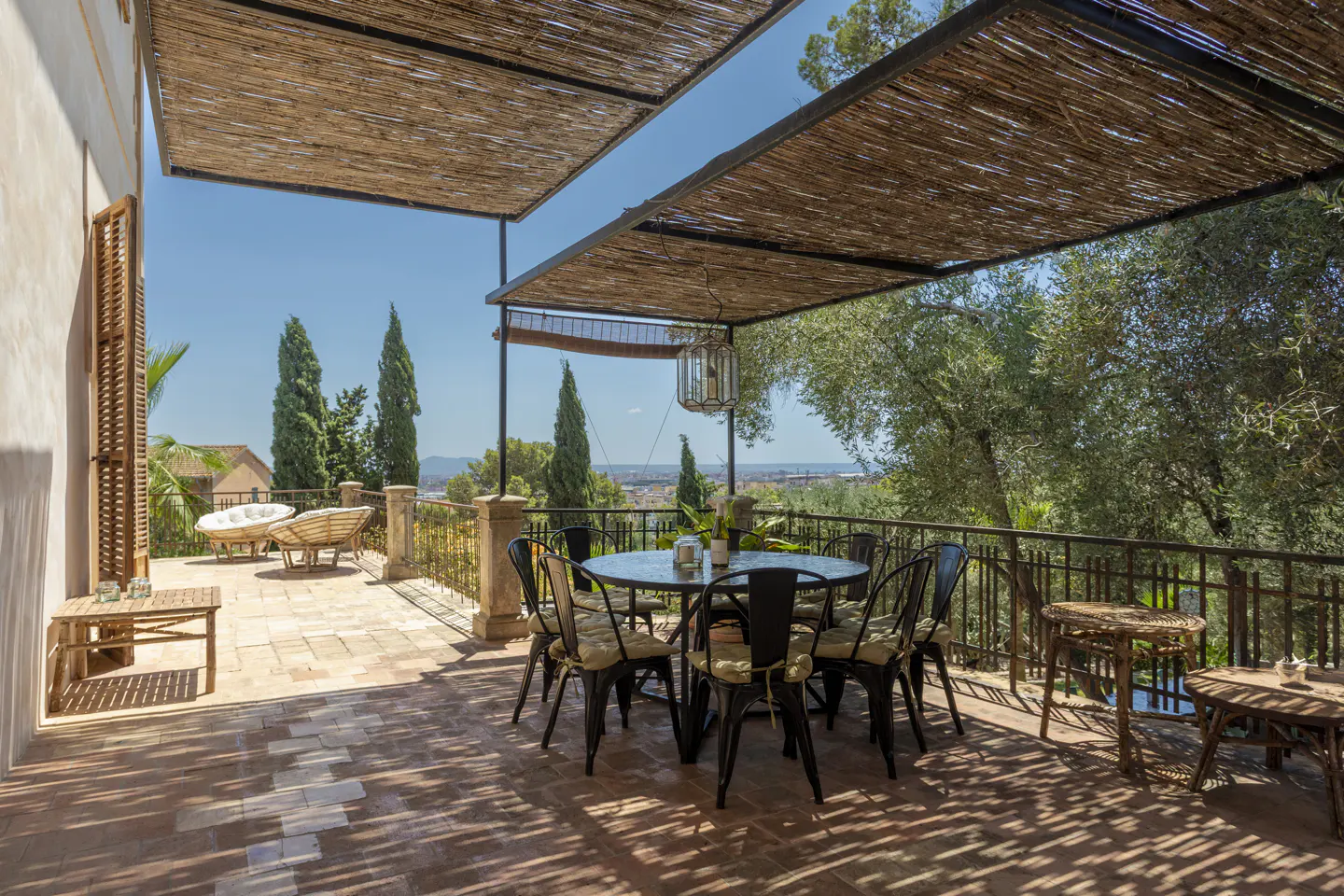 Outdoor patio with a round table, black chairs, and a straw roof. Trees and a city view are in the background.