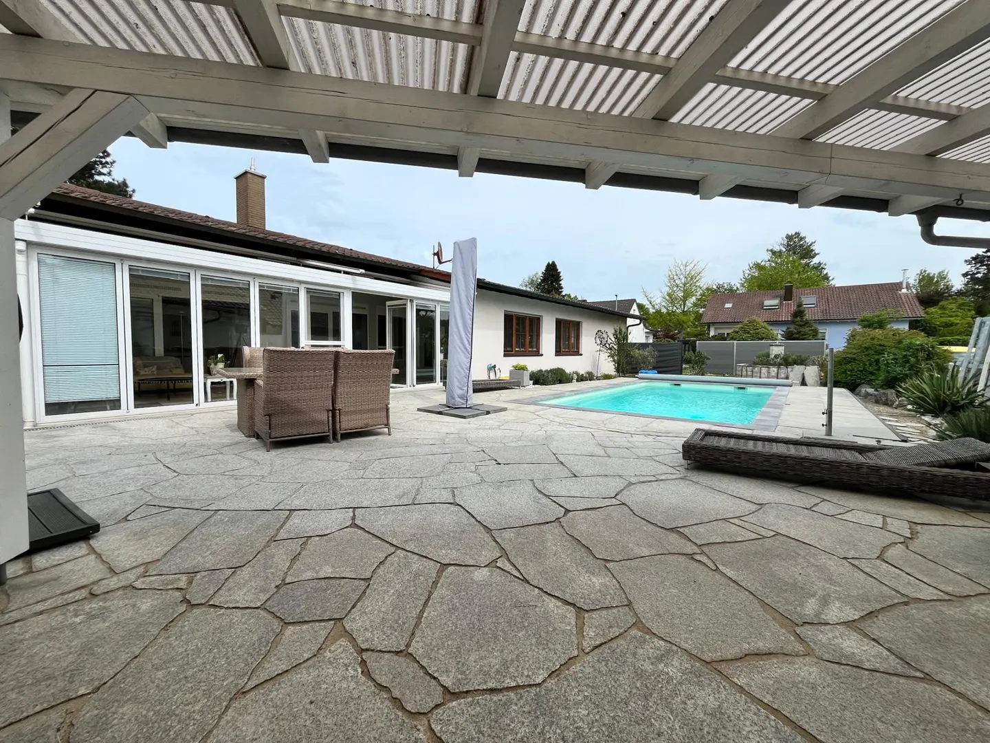 Outdoor patio with stone flooring, wicker furniture, and a pool. The house is white with brown trim. The sky is overcast.