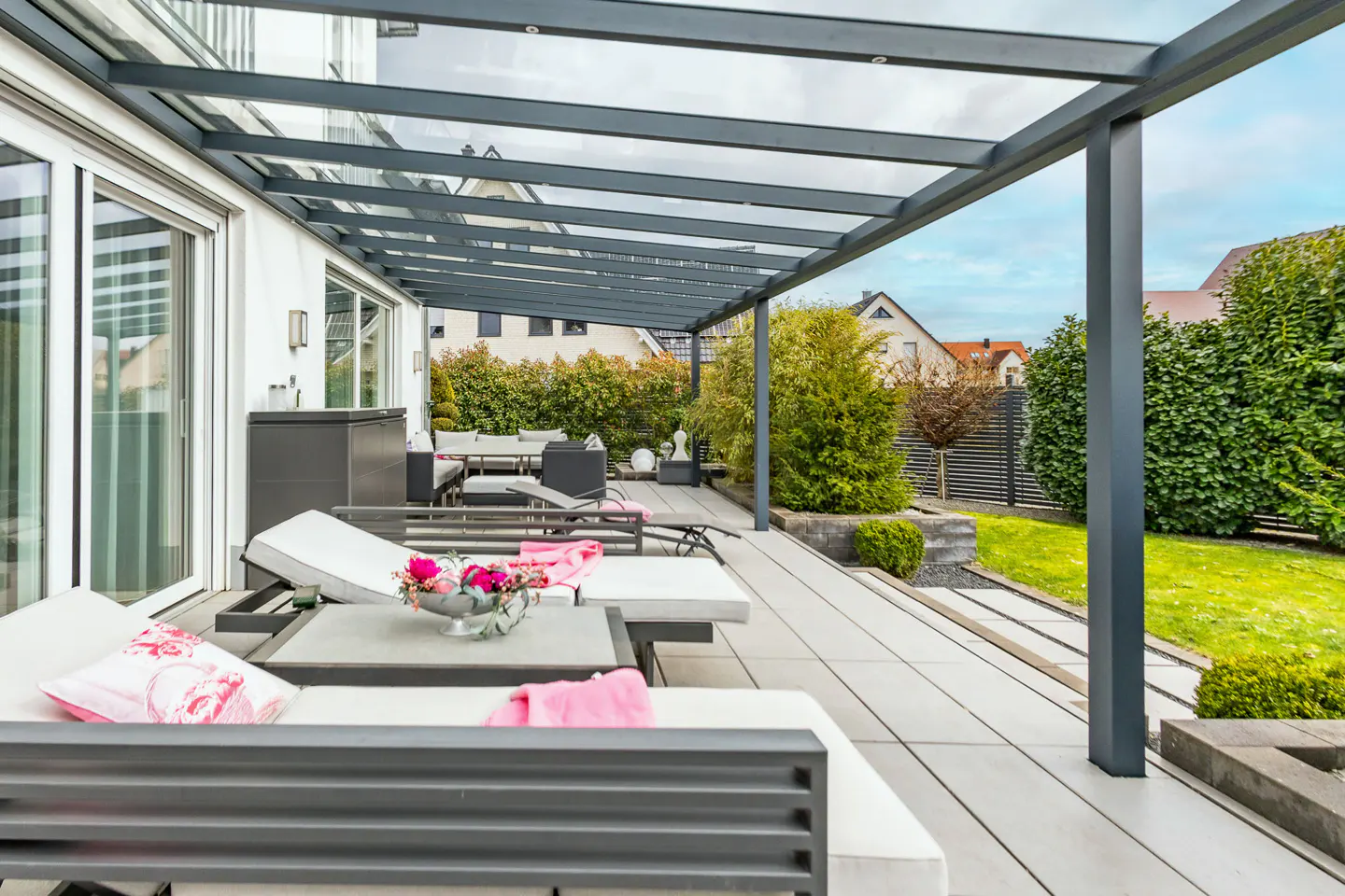 A modern patio with gray metal pergola, white lounge chairs with pink accents, and a lush green lawn.