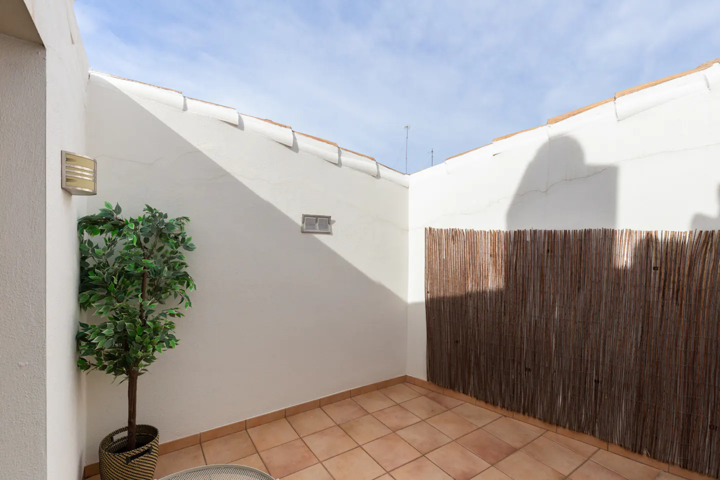 A small, private outdoor patio with terracotta tile flooring, white walls, a potted green plant, and a bamboo privacy screen.