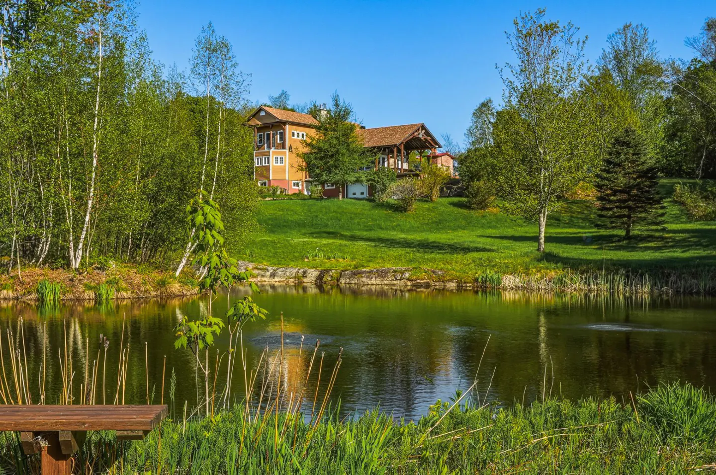 A yellow house sits on a green hill overlooking a pond, surrounded by trees under a blue sky.