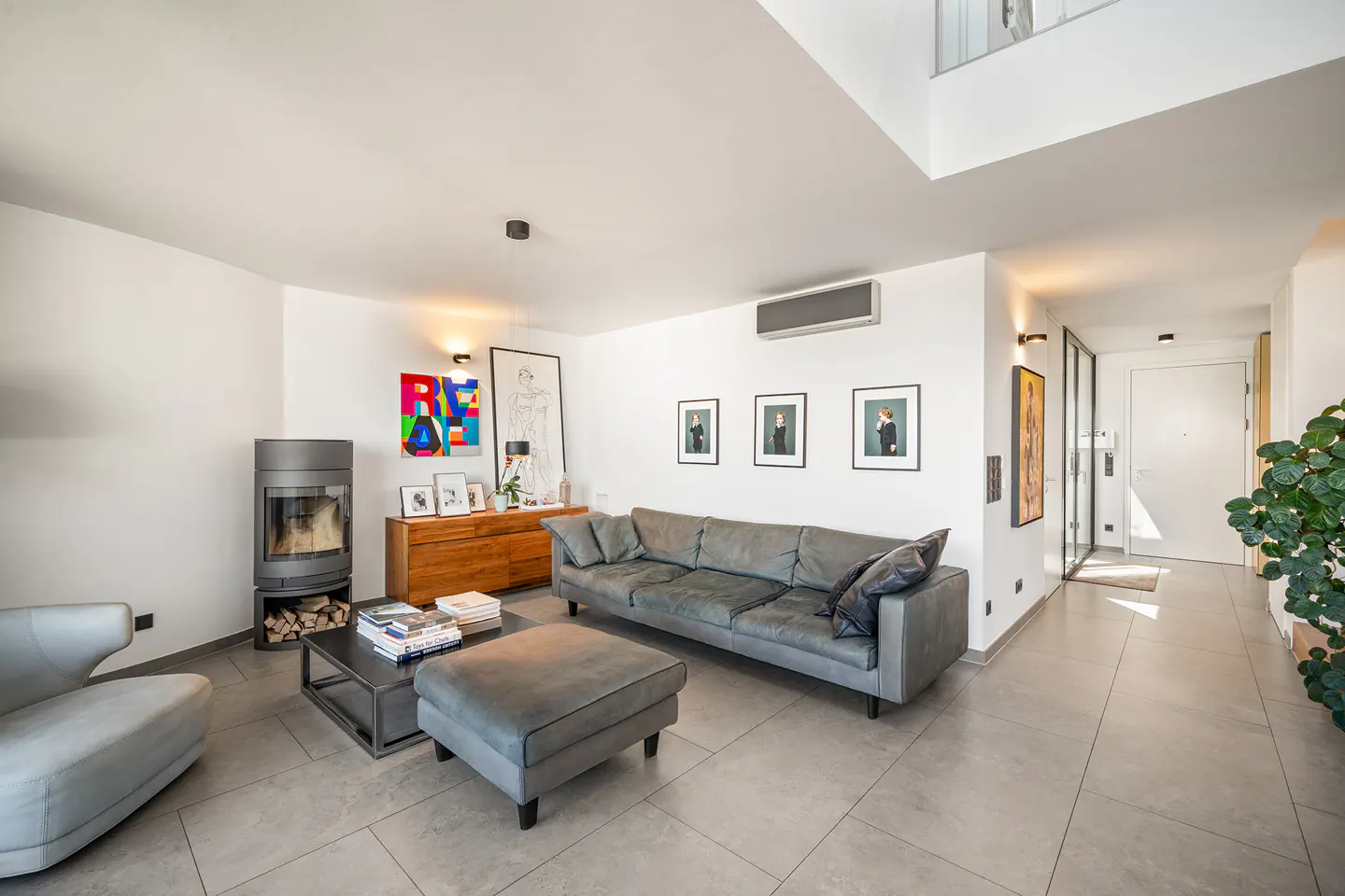 Modern living room with grey sofa, ottoman, wood-burning stove, and colorful wall art on white walls with tile flooring.
