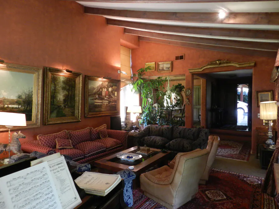 Living room with red walls, wooden beams, and paintings. Sofas, chairs, and a coffee table furnish the space. Sheet music sits on a piano.