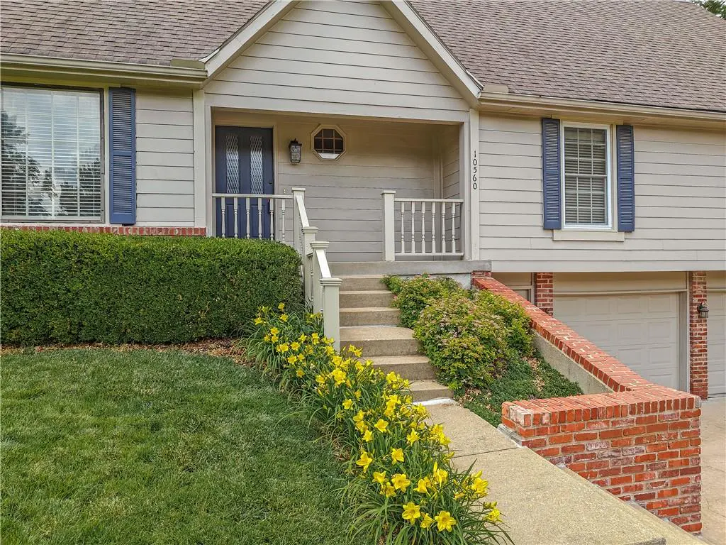 Beige two-story house with blue shutters and door, concrete steps, yellow flowers, and a green lawn.