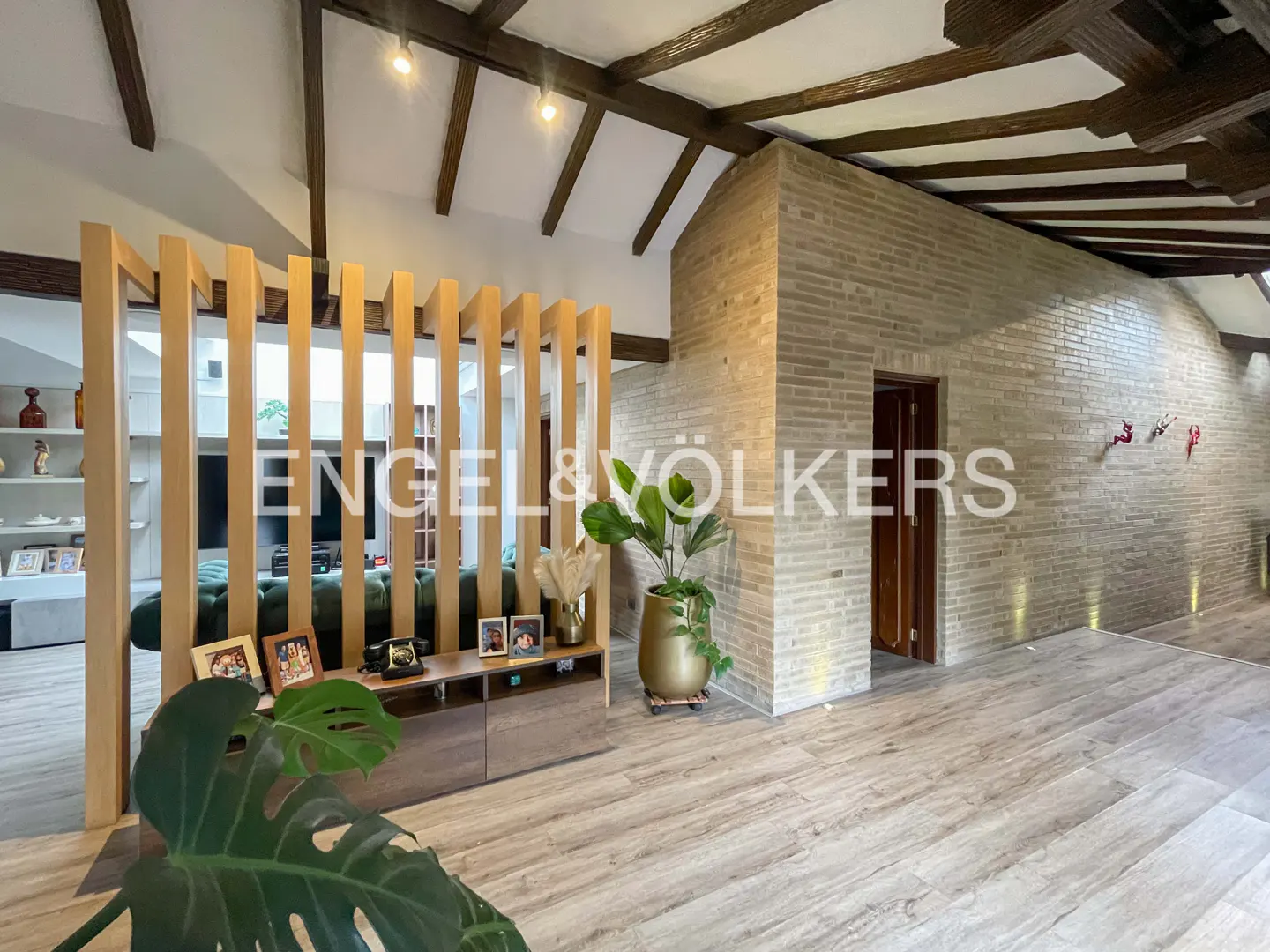 A modern living room with wood floors, exposed beams, and a brick wall. A wooden screen divides the space. A potted plant sits near a console table.