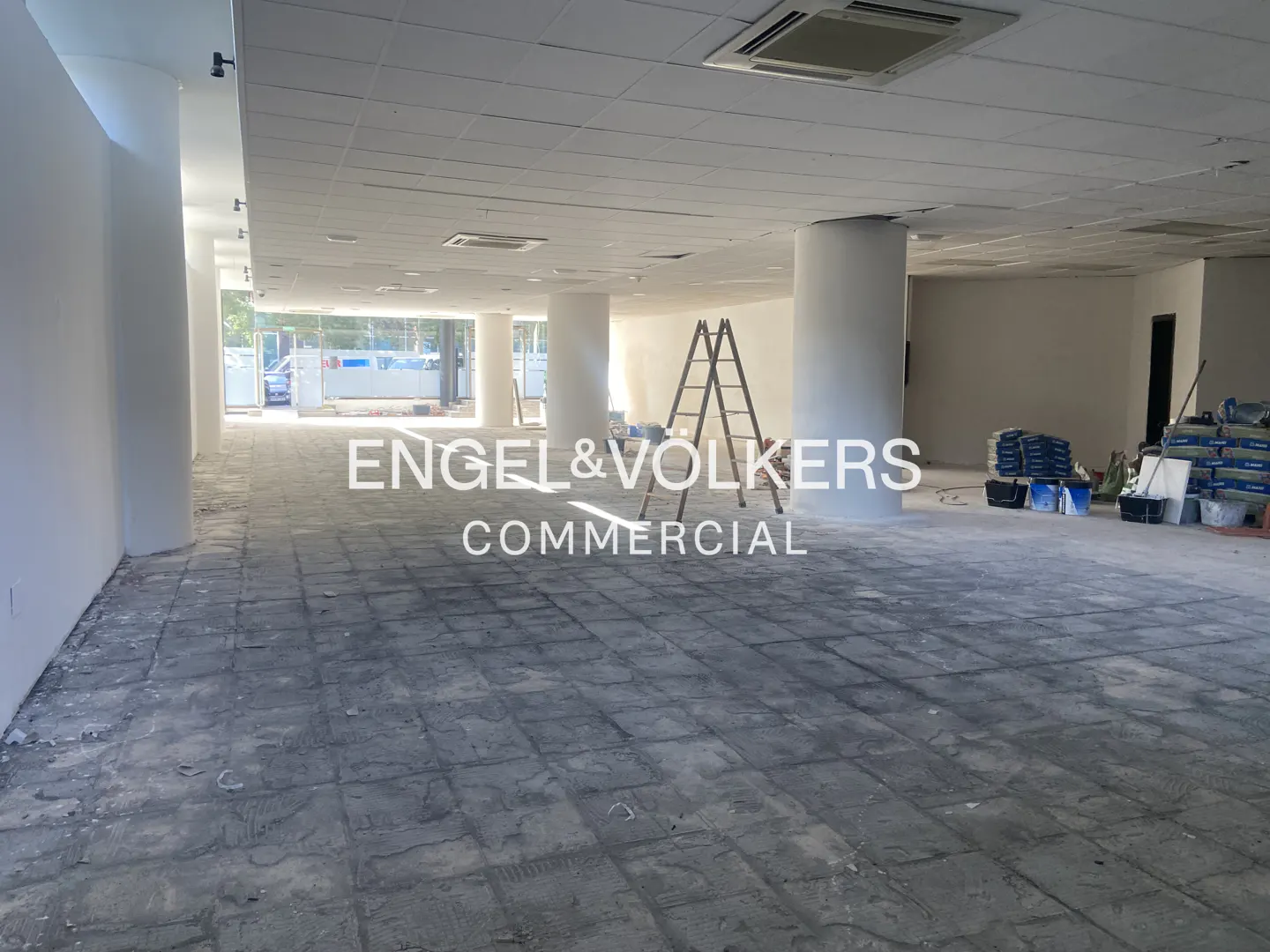 An empty commercial space with white walls and pillars, a ladder, and construction materials. The floor is gray and unfinished.