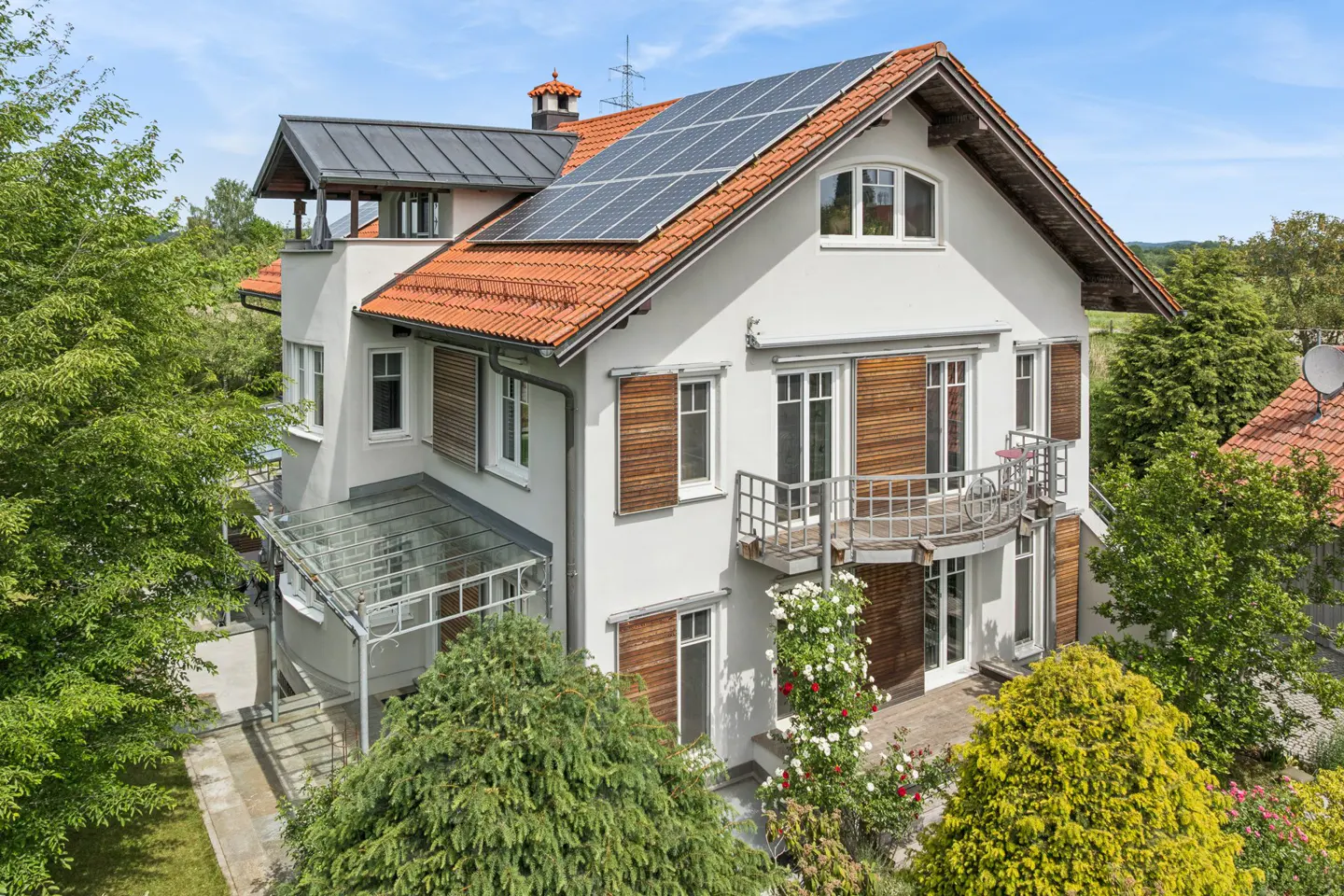 White house with orange tile roof, solar panels, wooden shutters, a curved balcony, and a glass-covered patio in a garden.
