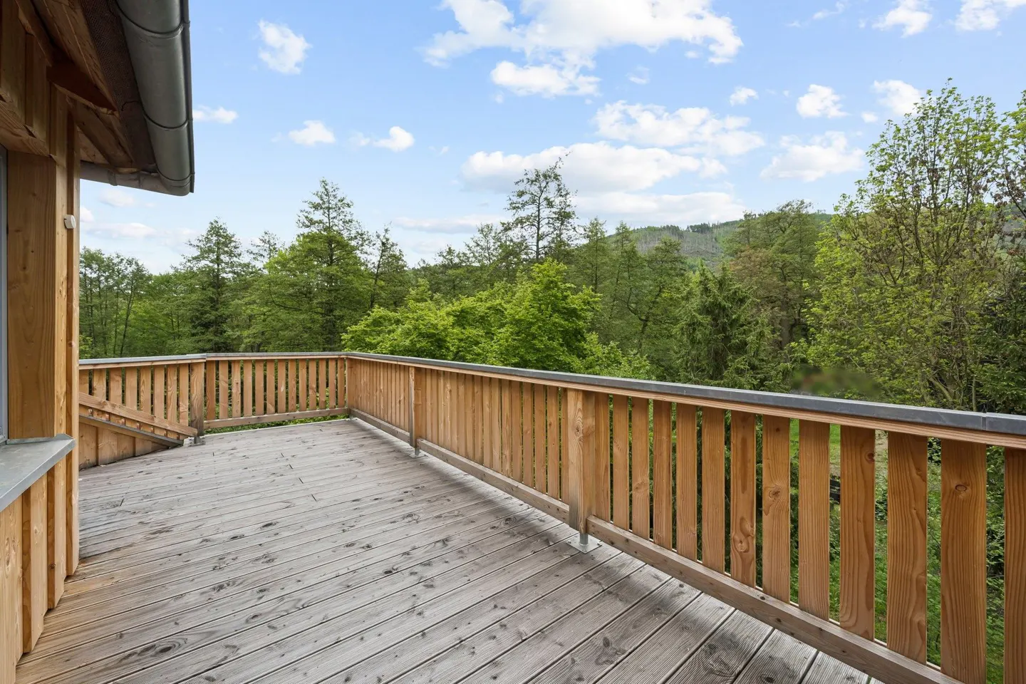 Wooden deck with a railing overlooks a lush green forest under a blue sky with scattered clouds.